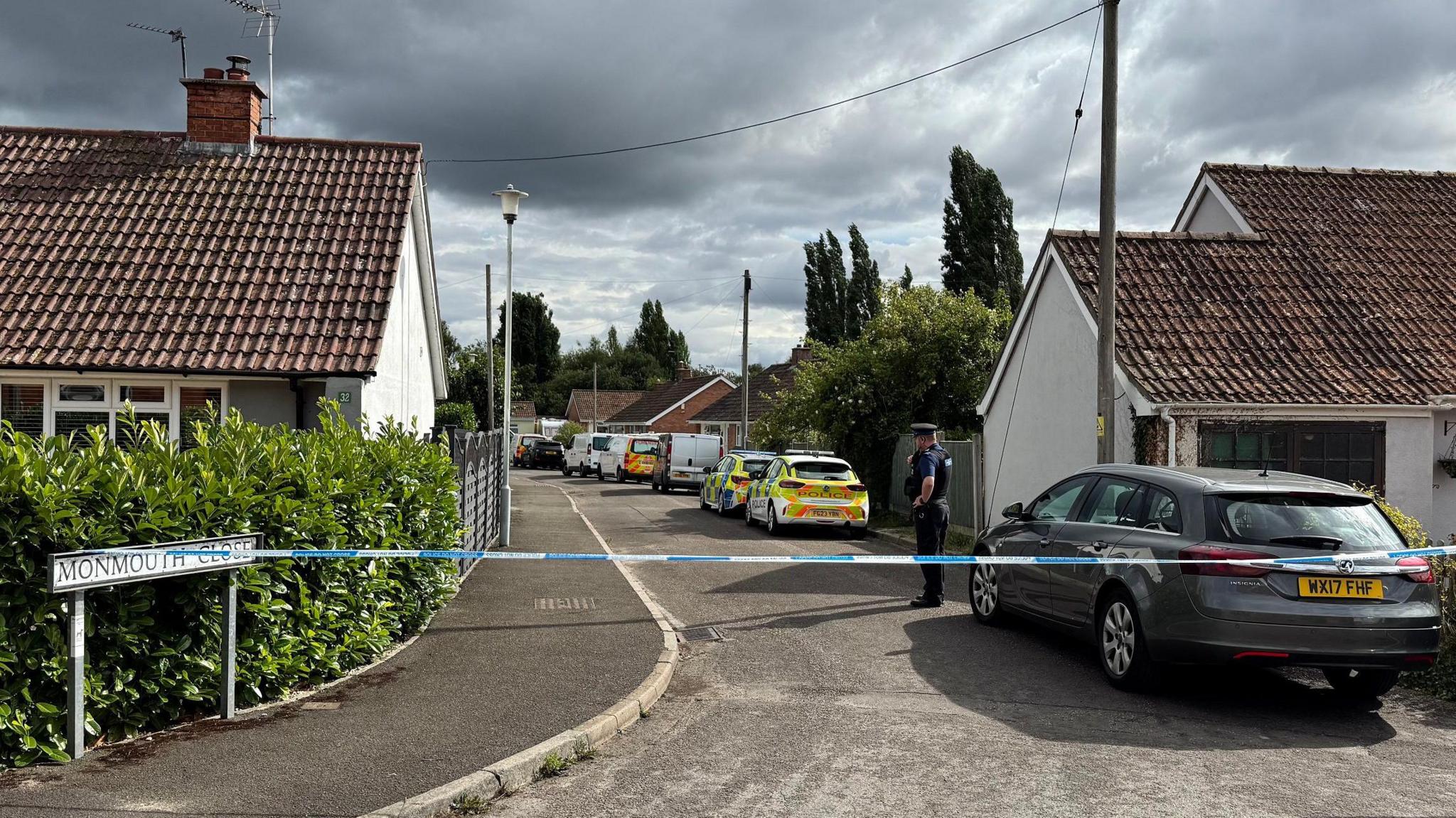 A general view of Monmouth Close in Westonzoyland, which has been fully cordoned off with blue and white police tape. The street is made up of a small number of bungalows with red tile roofs. A number of police cars and vans can be seen parked on the road and a police officer is shown next to the cordon.