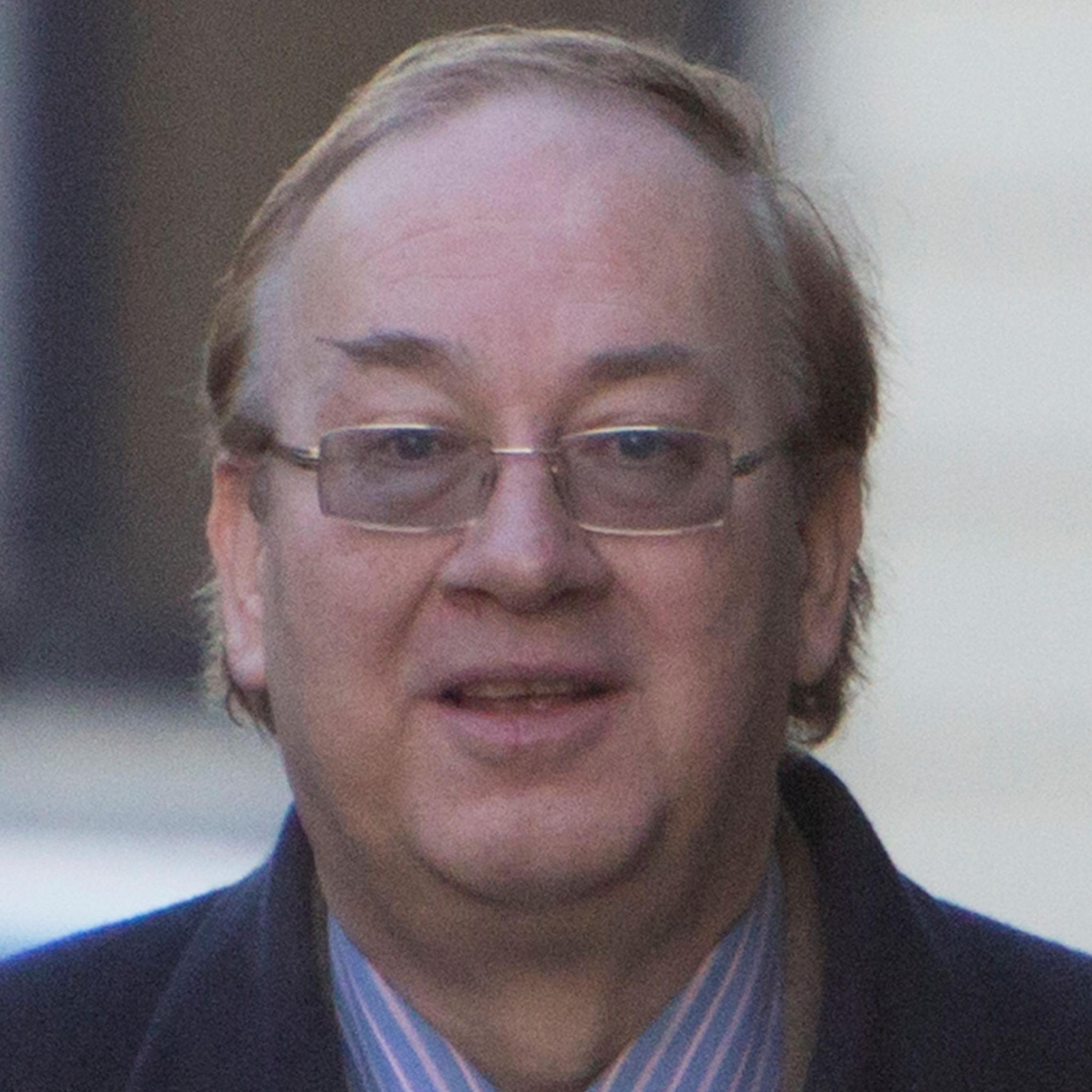 Kevin Booth is looking at the camera. He is wearing a blue jacket over a suit and stripy shirt. He is wearing glasses.