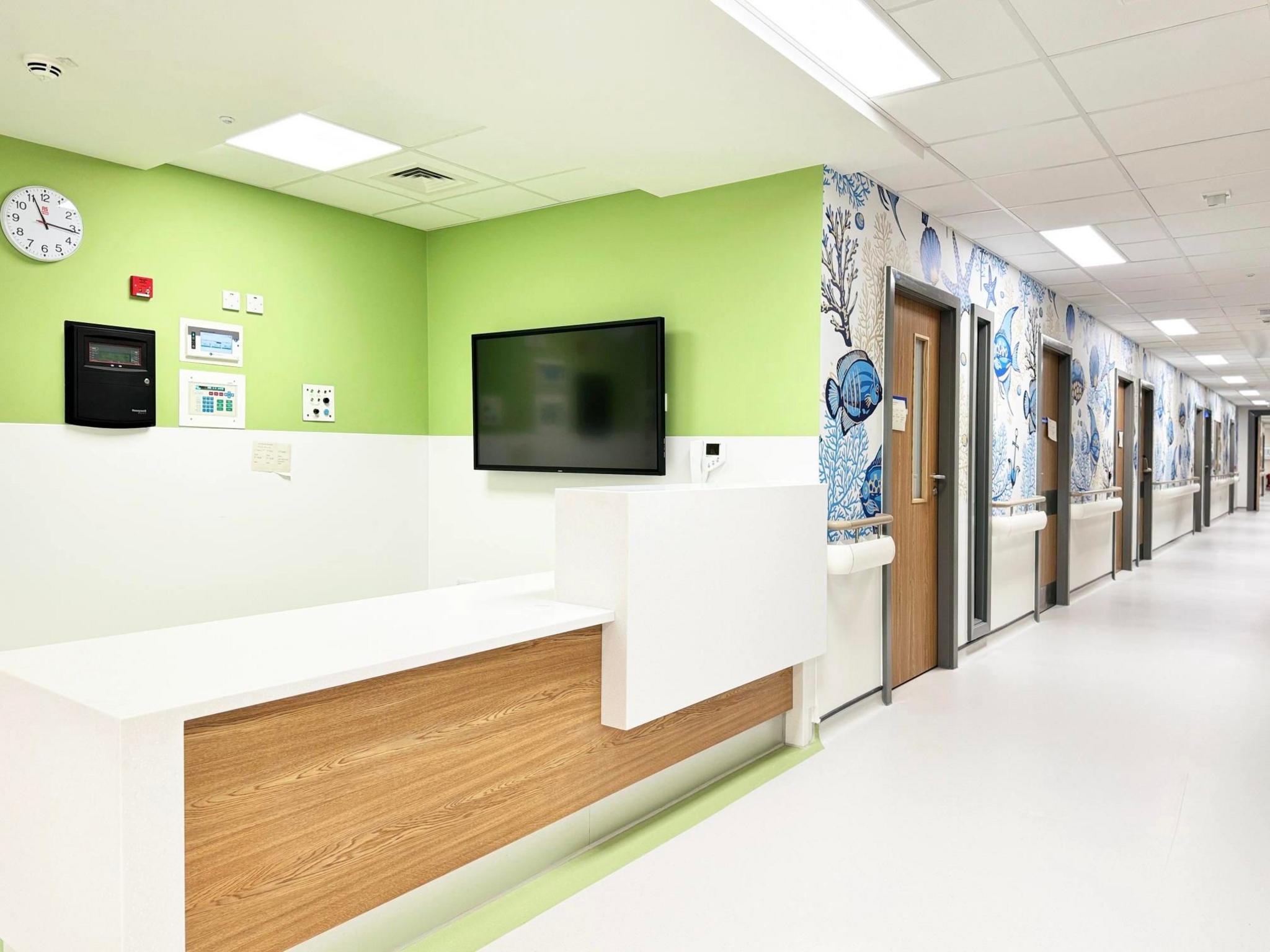 The children and young people's ward has light green and blue, patterned walls. A reception desk is seen in the foreground of the image, with a corridor in the background.