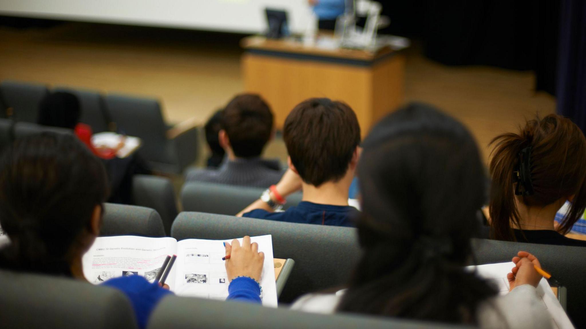 Students in a lecture theatre. The students, all facing away from the camera, are taking notes in notepads while a lecturer, wearing a blue shirt, stands at the front of the hall in front of a wooden desk.