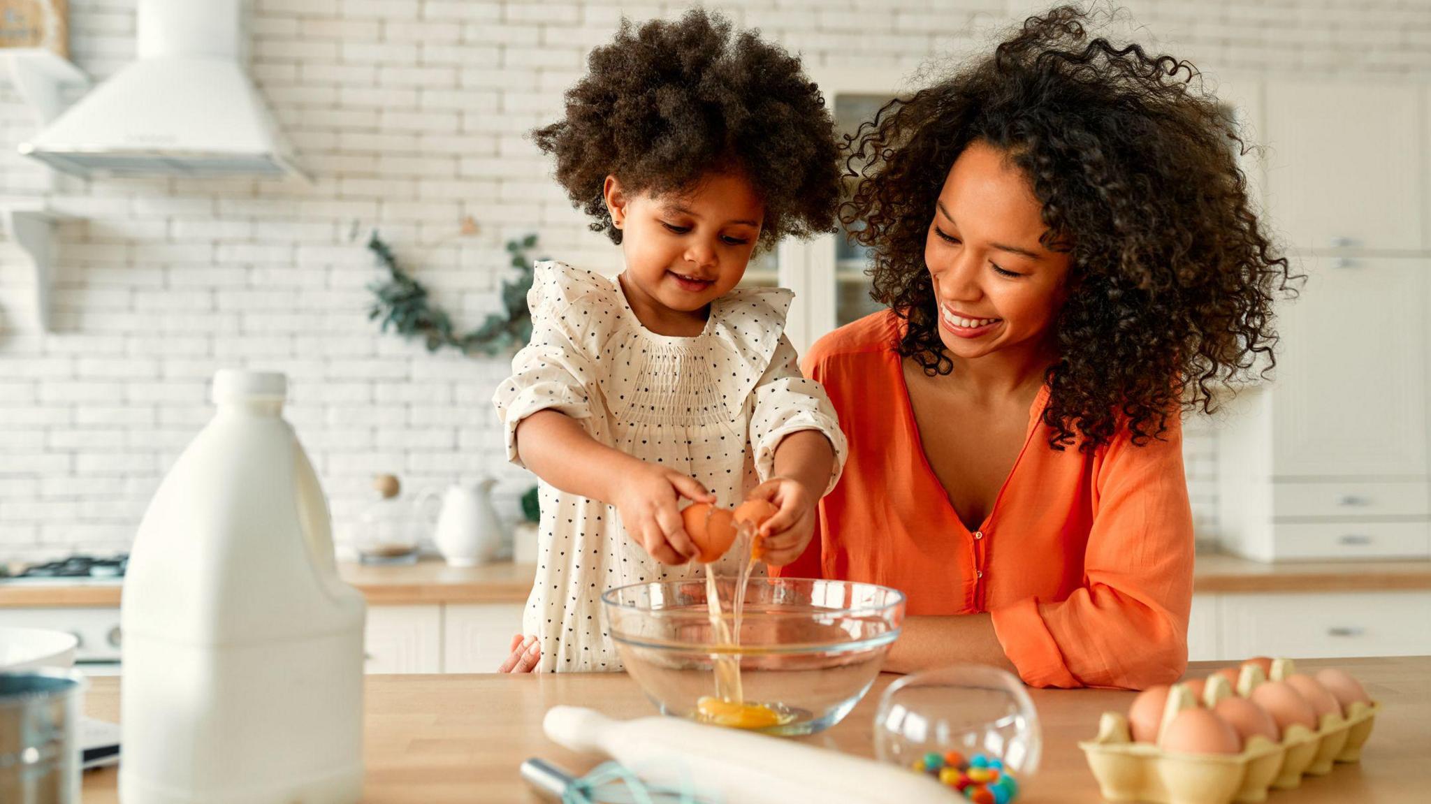 A child and her mother sat on a kitchen table, the child wears a white polka dot dress and the mother wears a bright orange blouse, the child is cracking an egg into a bowl.