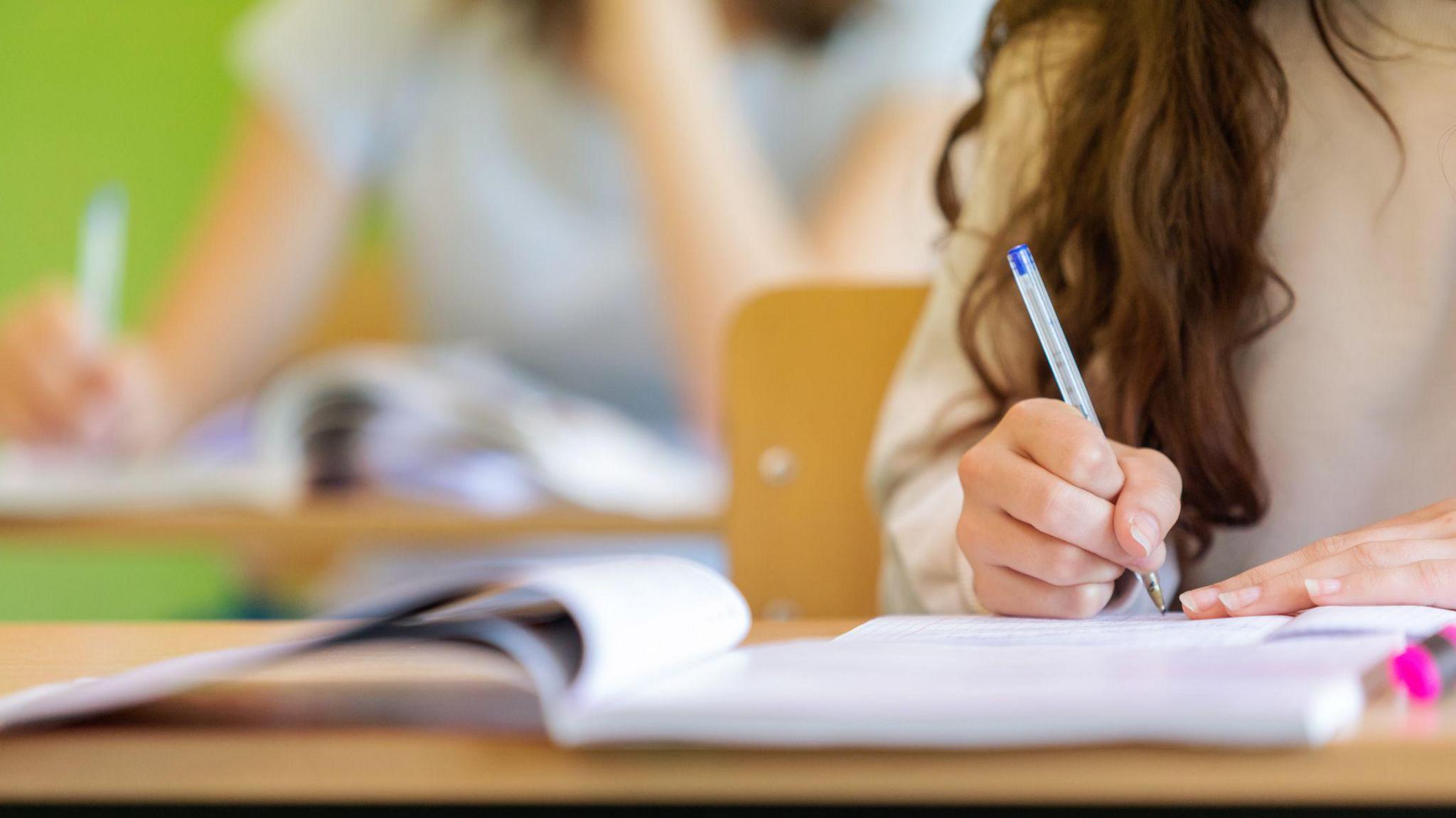 A student sitting at a desk, with long brown hair, writes with a blue pen on a piece of paper. The student's face is not in view.