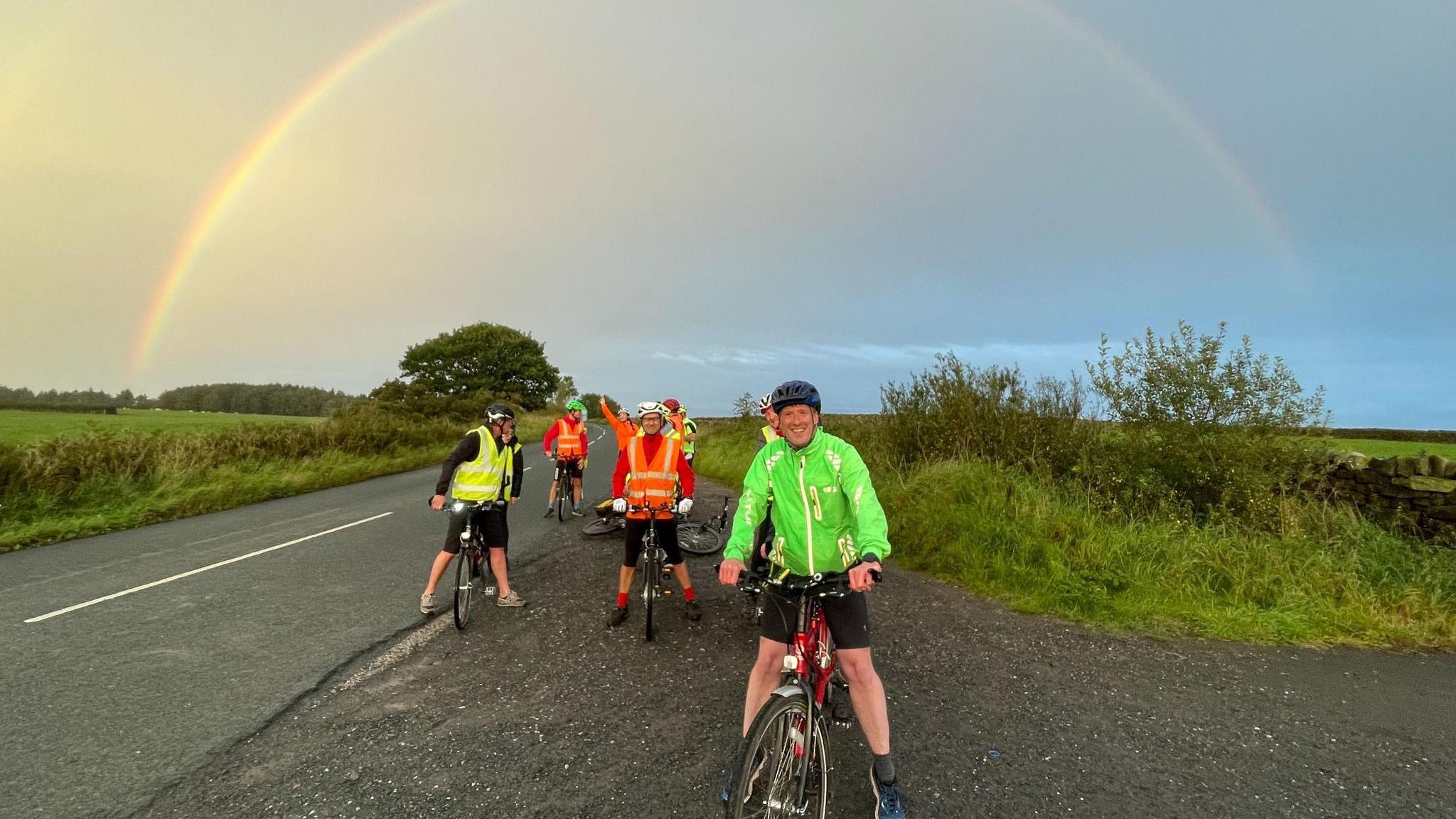 A group of people on tandem bikes, wearing reflective cycling gear and helmets pose by the side of a road, next to a field, under a rainbow in the sky.