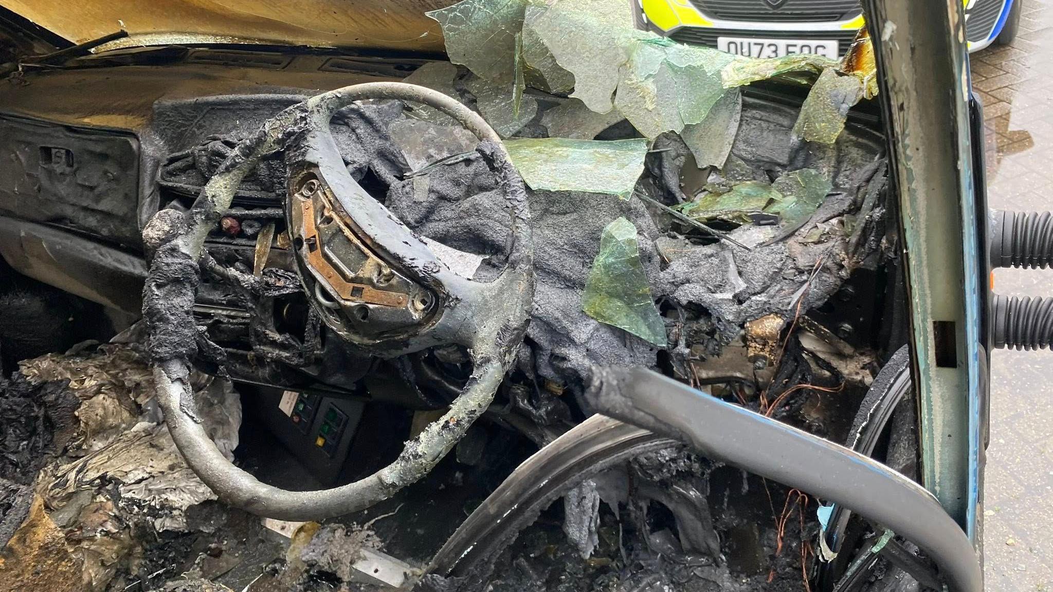The inside of a van which is melted and charred. The windscreen is smashed.