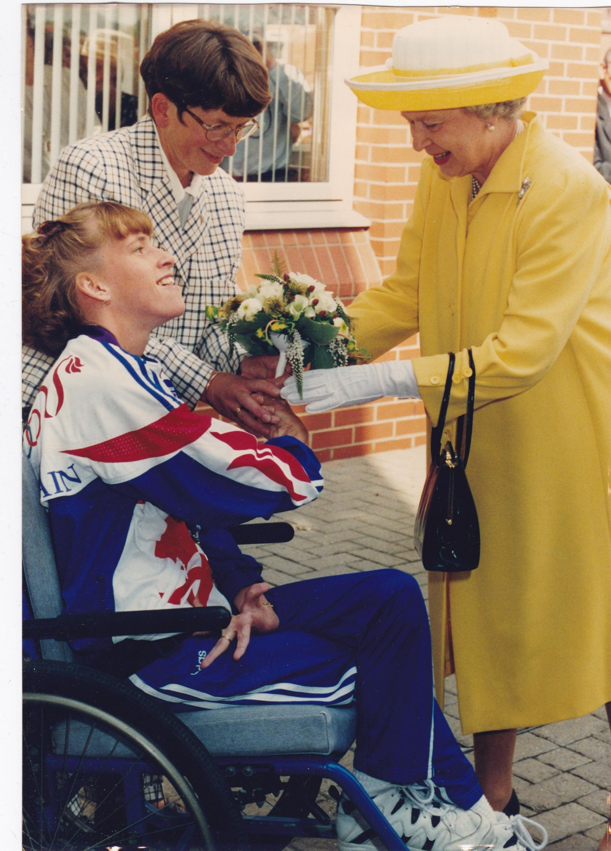 A young woman in a tracksuit hands flowers to the Queen dressed in a yellow coat and white gloves