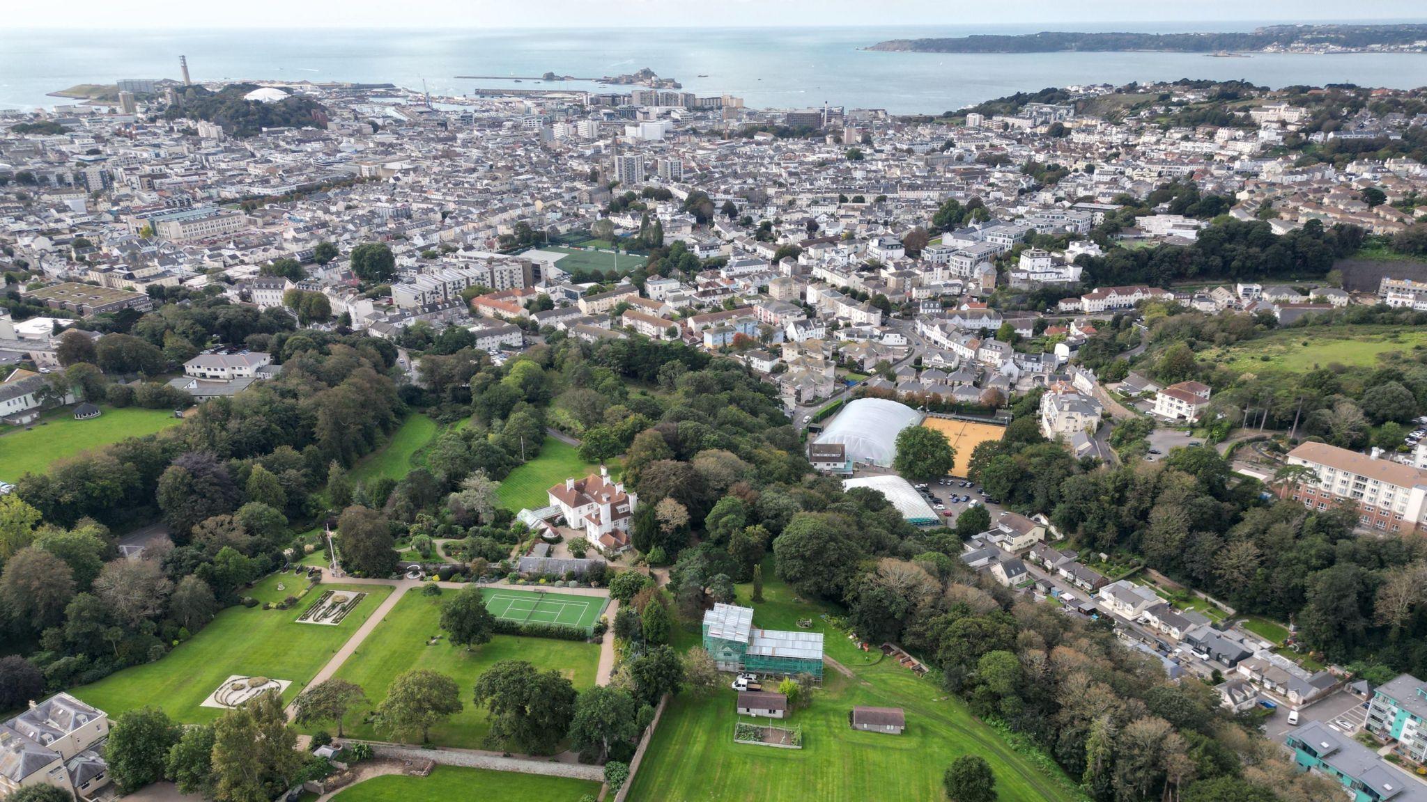 A drone photo of St Helier and some of Jersey. In the distance it shows the sea and then shows buildings. There is also a large site of green as well as trees.