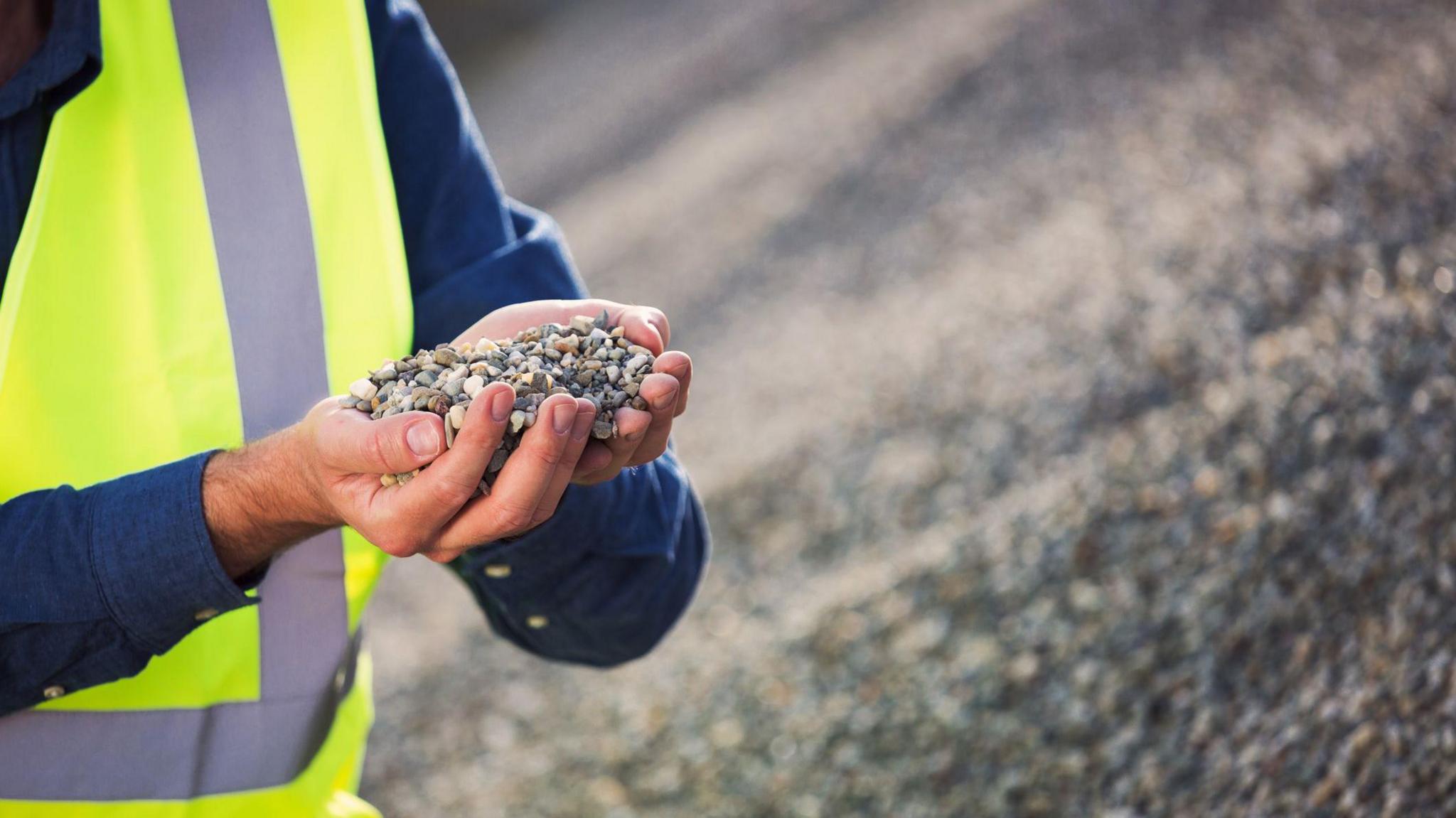 A man in a blue shirt covered with a hi-viz jacket holds up some gravel in his hands, behind him is a pile of gravel