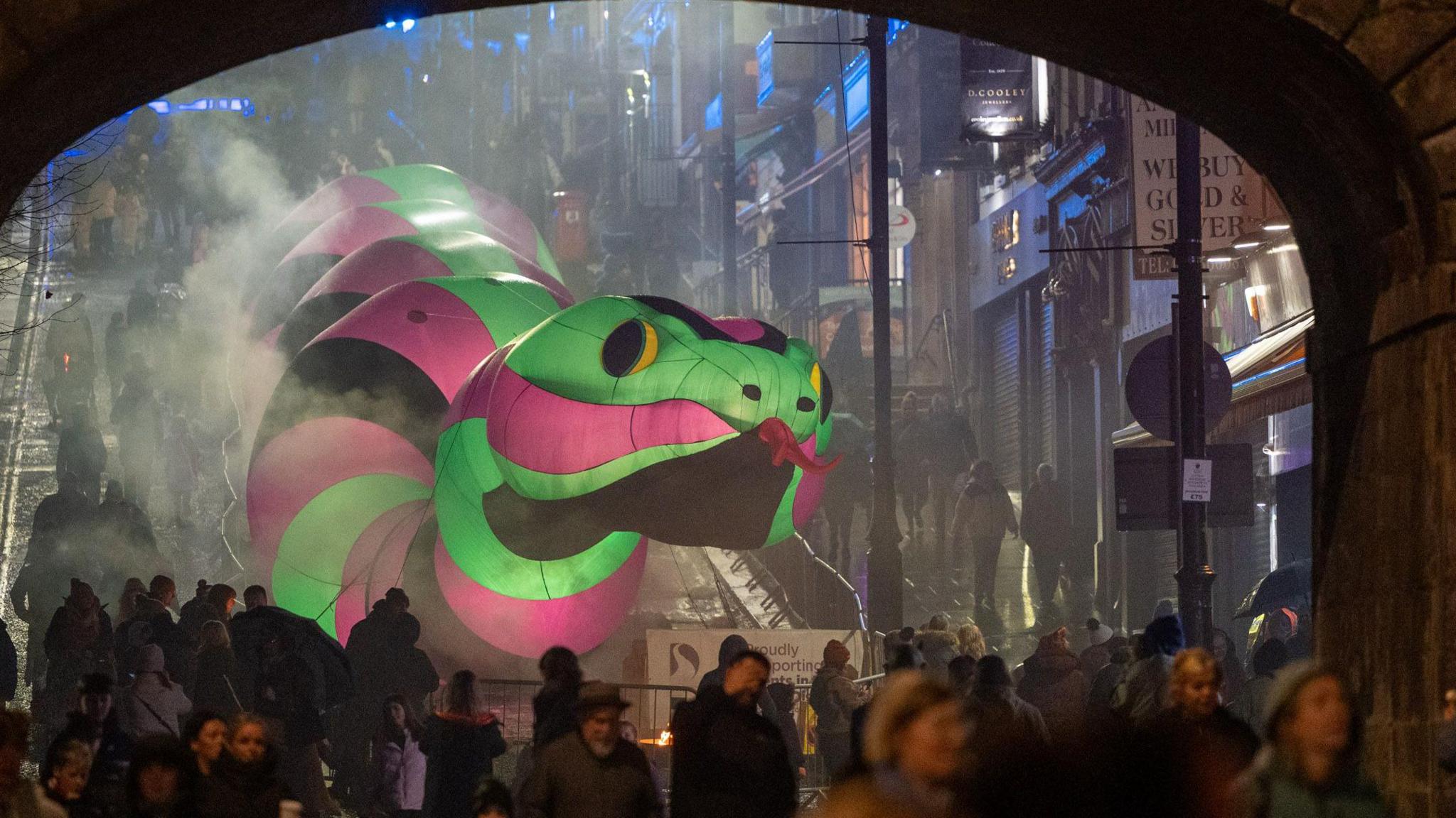 Shows a large illuminated snake in green, pink and black going down Shipquay Street, with people in the foreground in front of an arch.