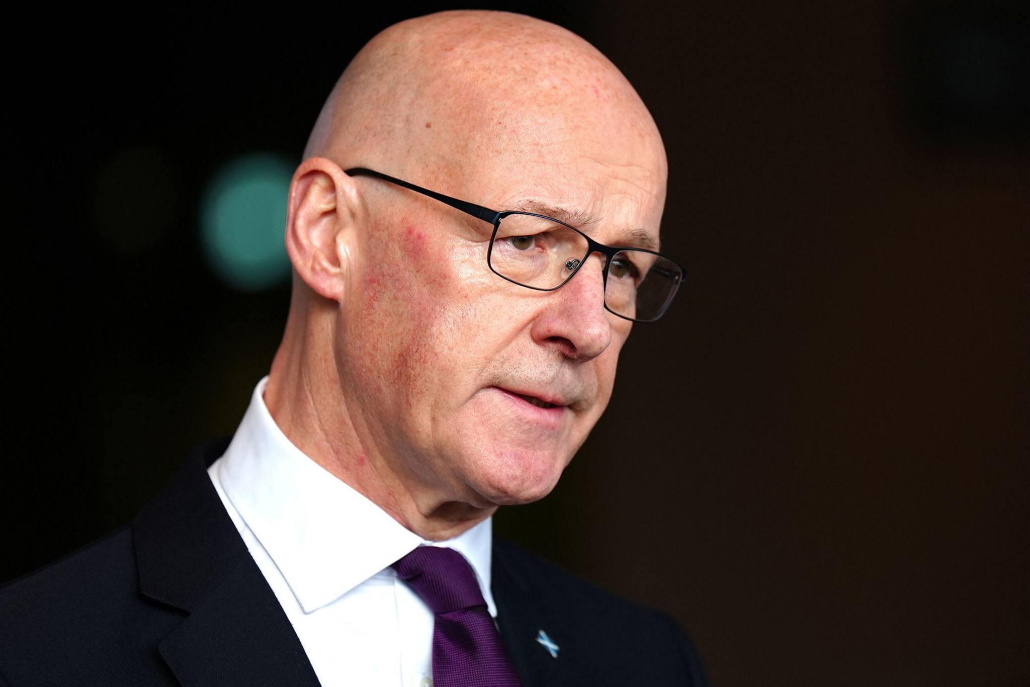 A bald headed man with glasses, wearing a purple tie, white shirt and dark suit