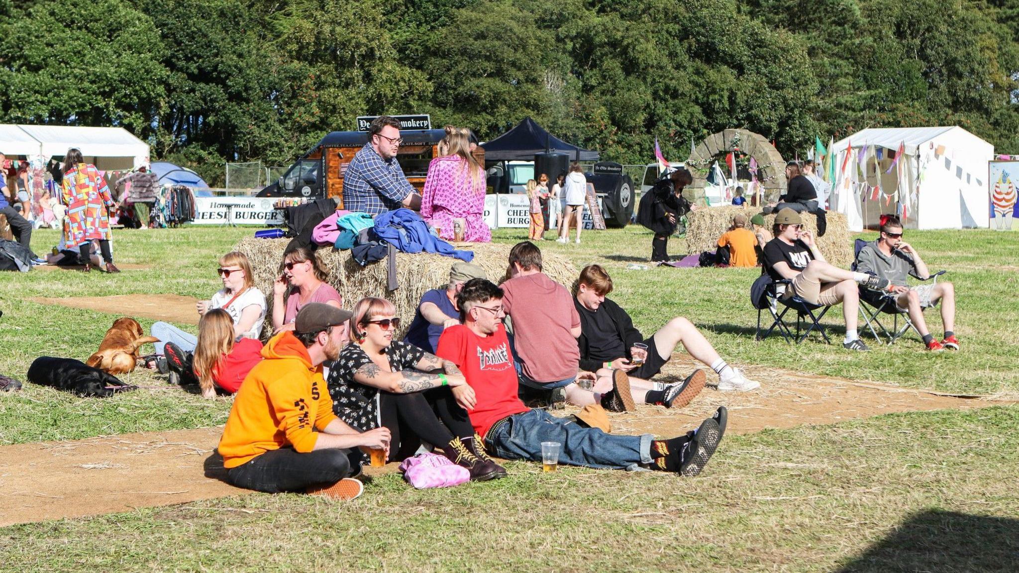 Festival goers sitting on grass, enjoying the sunshine. Several stalls and gazebos can be seen in the background.