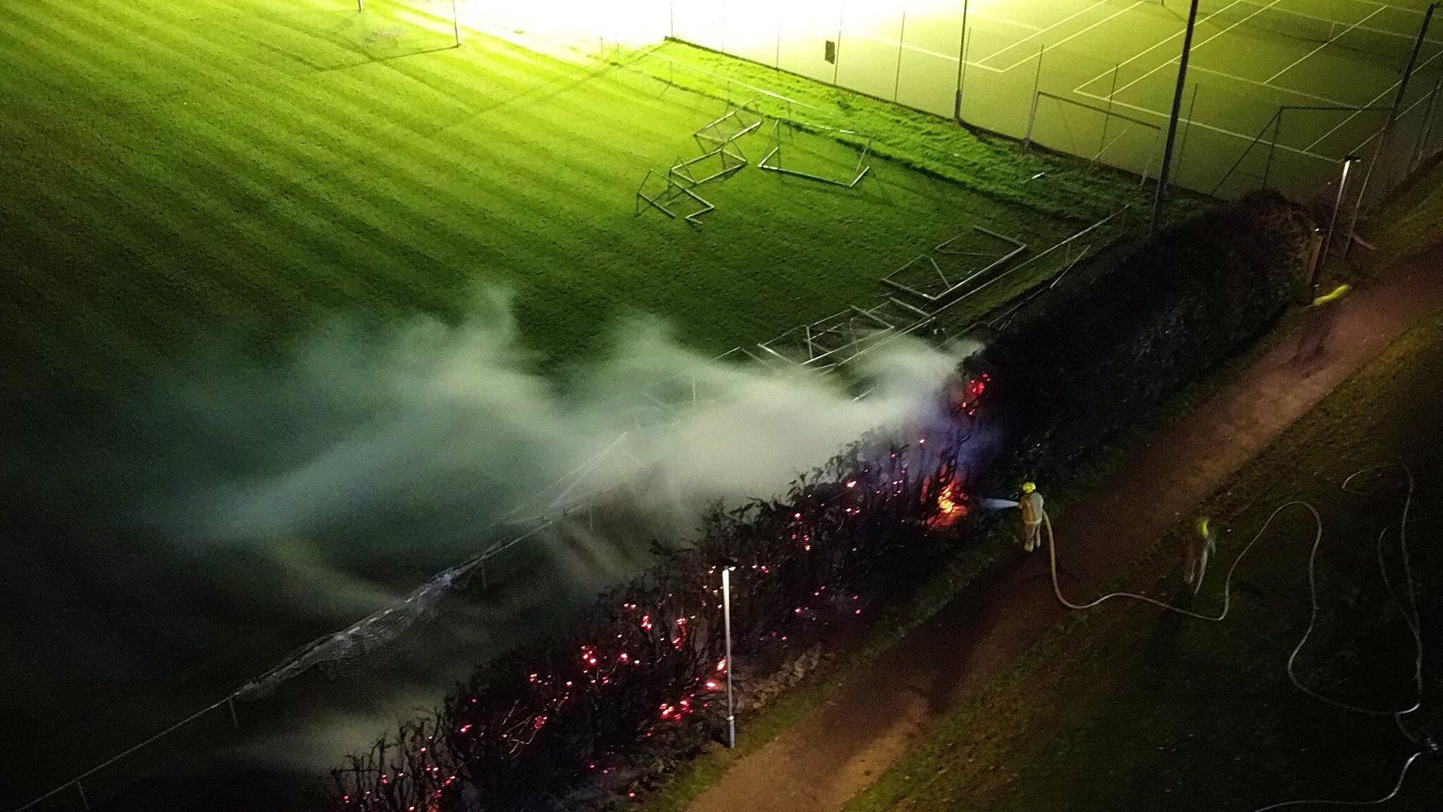 Flames and smoke coming from hedges surrounding football pitches. The sky is dark and firefighters and members of the public are standing nearby. Floodlights are lighting up the area. 