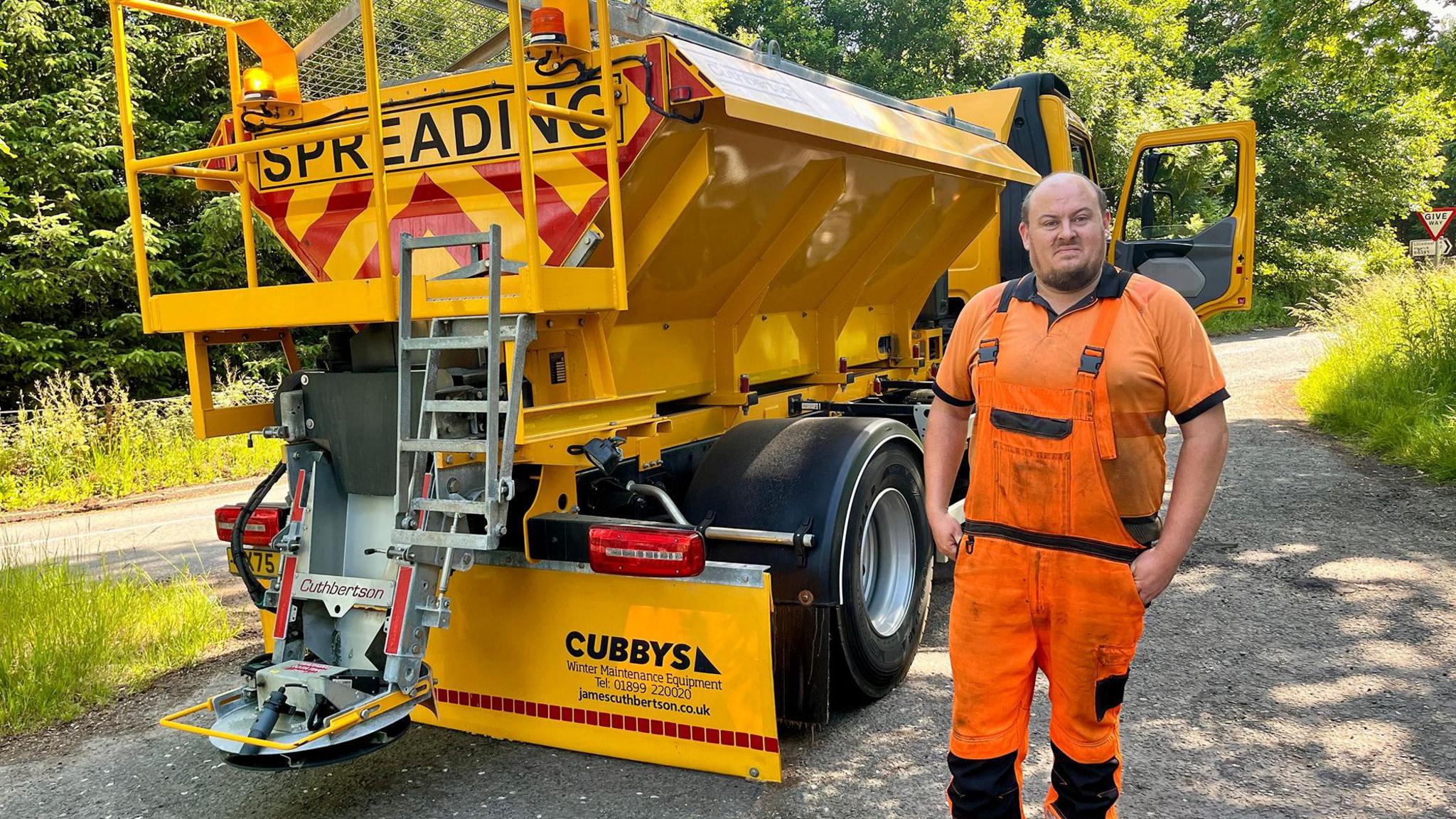 Road worker in orange high-visibility clothing standing beside a yellow gritting truck marked Spreading on a rural roadside with trees in the background.