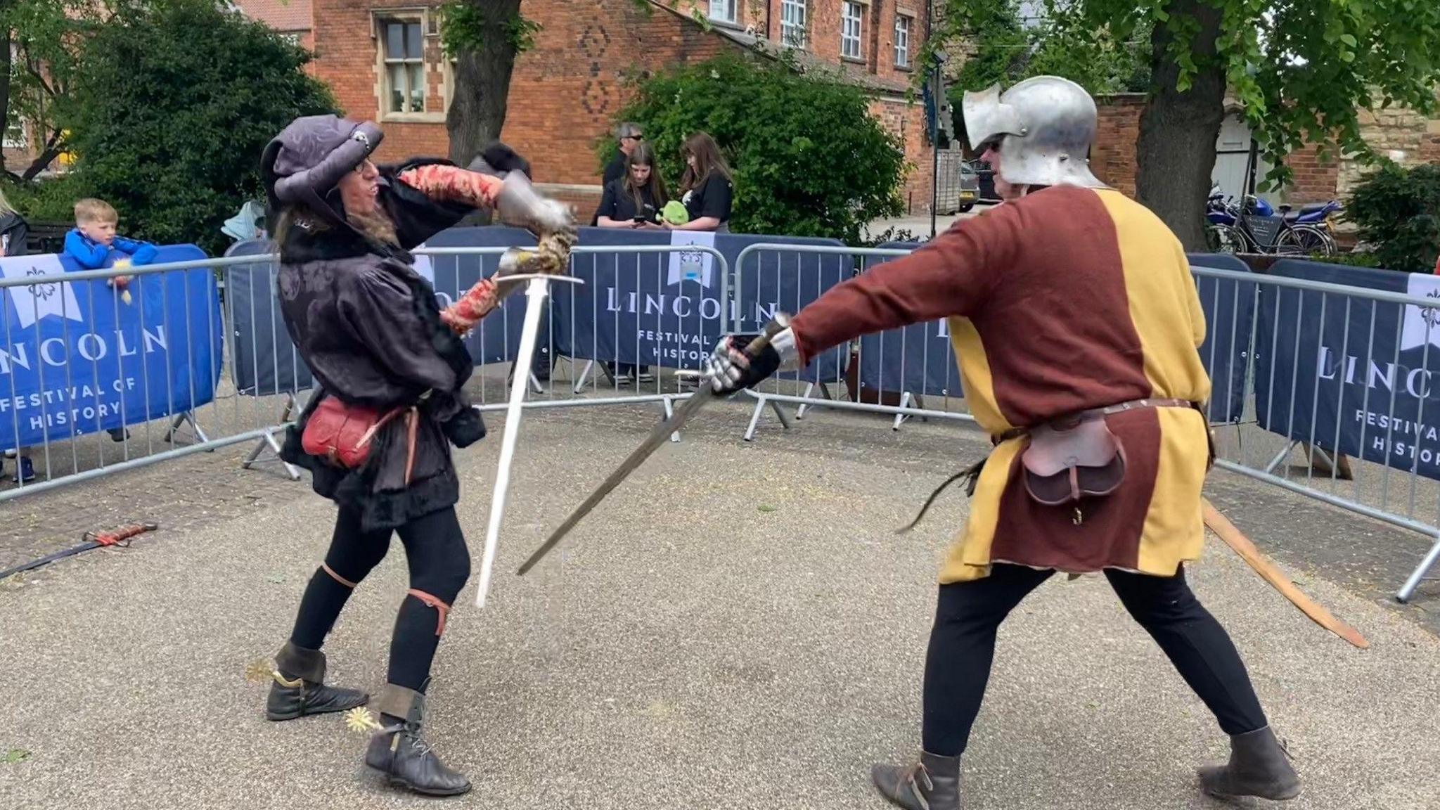 Two men in medieval costumes fight with swords within a fenced off area.