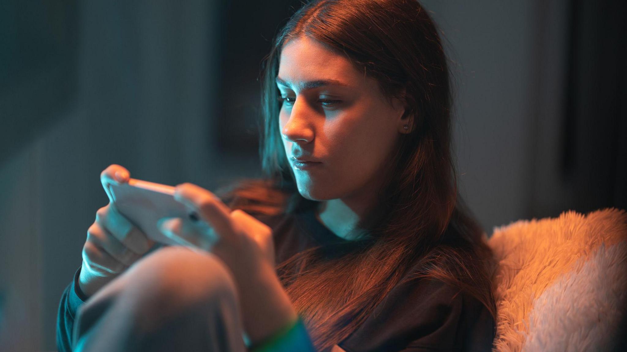 A young woman plays a mobile game on her phone in a dimly-lit room