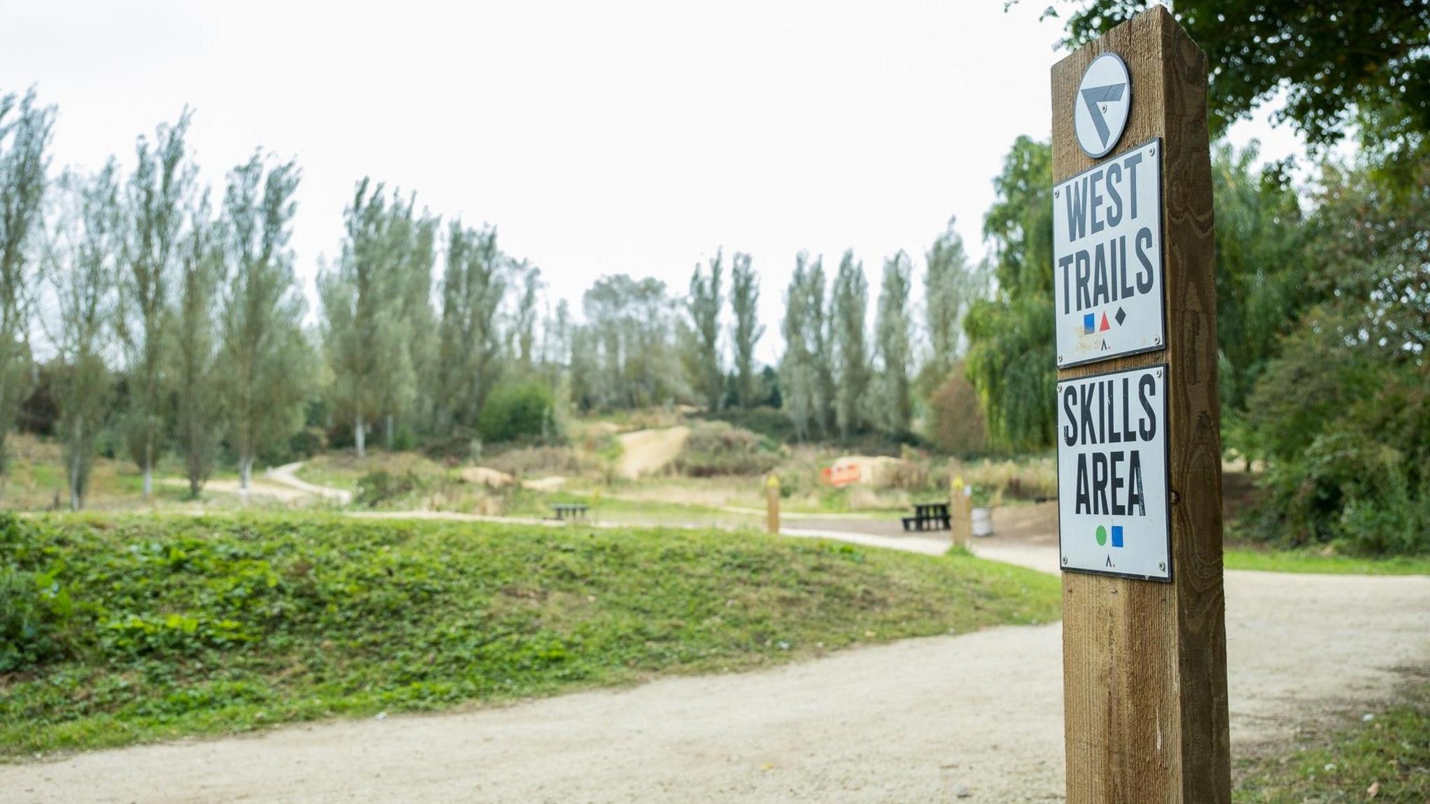 A cycle trail making its way through grass hills. A line of trees is visible in the background and a wooden post in the foreground has signs saying "West Trails" and "Skills Area".