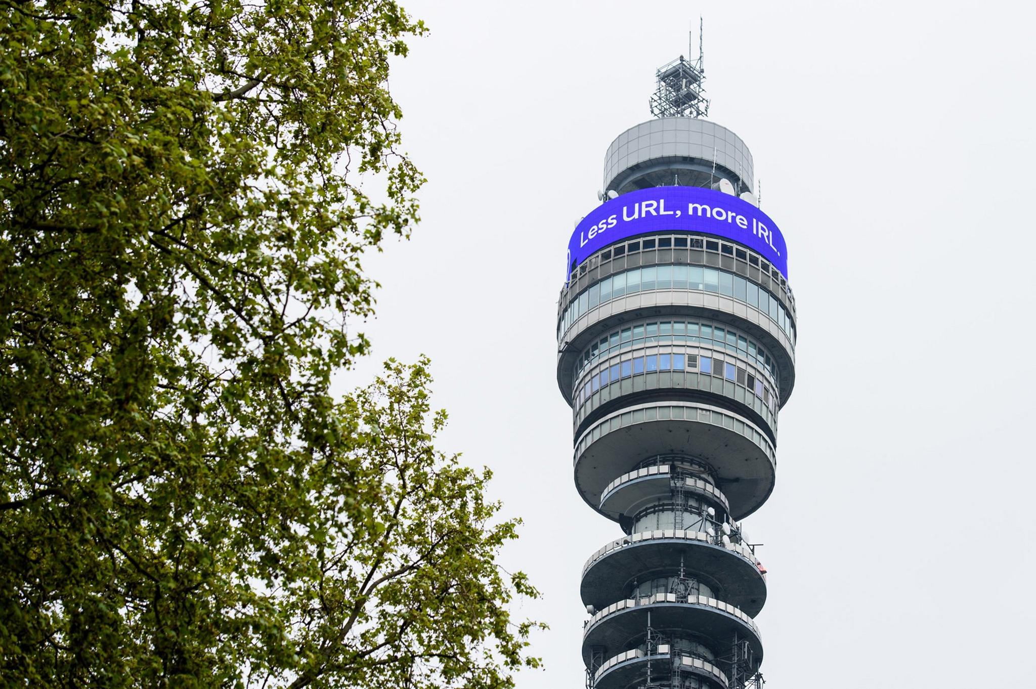 A digital message, which reads Less URL, more IRL. Welcome back is displayed on the BT Tower above the streets of London