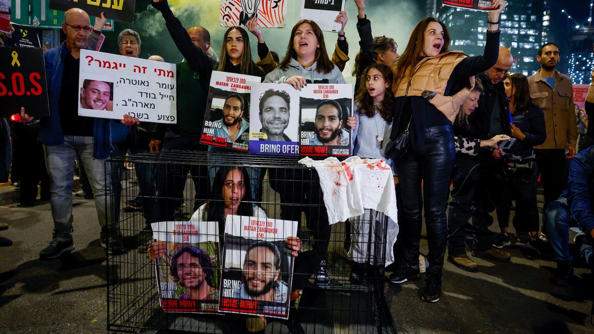 A woman sitting in a cage holds posters showing two Israeli hostages still being held by Hamas in Gaza, at a protest in Tel Aviv, Israel (26 March 2024)