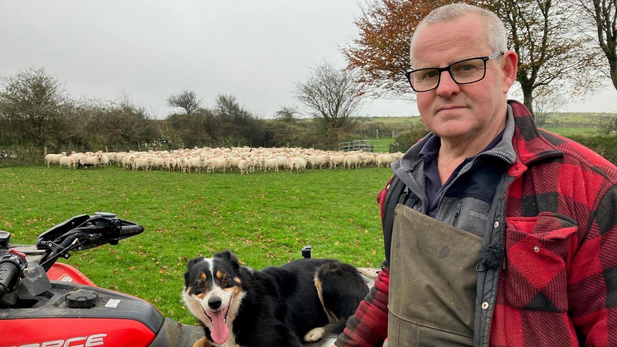 Farmer Colin Abel stands in the foreground wearing glasses, green overalls and a red and black checked overshirt. To his left is his sheepdog atop a red quad bike while in the background a flock of sheep can be seen in a field.