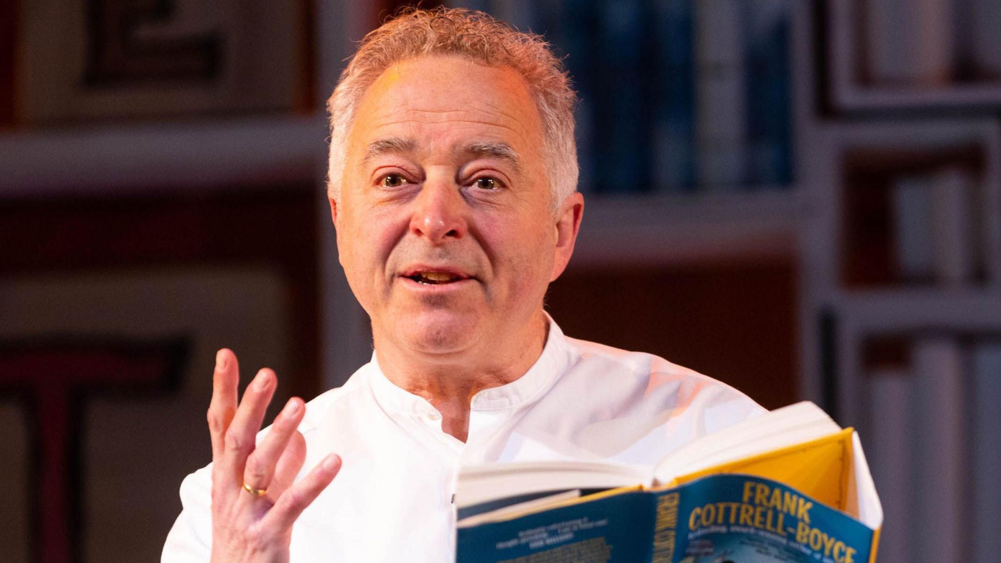 Frank Cottrell-Boyce on stage, gesturing with one hand and holding one of his books in the other