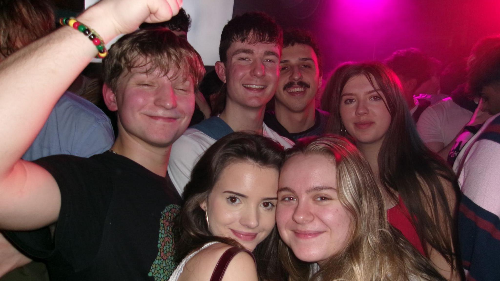 A crowded group photograph taken in a nightclub or live music venue. Several people are closely gathered together and filling most of the frame. Two people are positioned in the foreground, standing cheek-to-cheek near the centre, while additional people stand behind them in rows extending into the background. On the left side of the image, a raised arm wearing a multicoloured beaded bracelet reaches upward and partially enters the frame. The room is packed with people, with only small glimpses of walls and fixtures visible. Bright pink and purple club lighting shines from the upper right corner and reflects across faces, hair and clothing throughout the crowd. The setting appears busy and energetic, with people standing shoulder to shoulder in a lively indoor nightlife environment. 