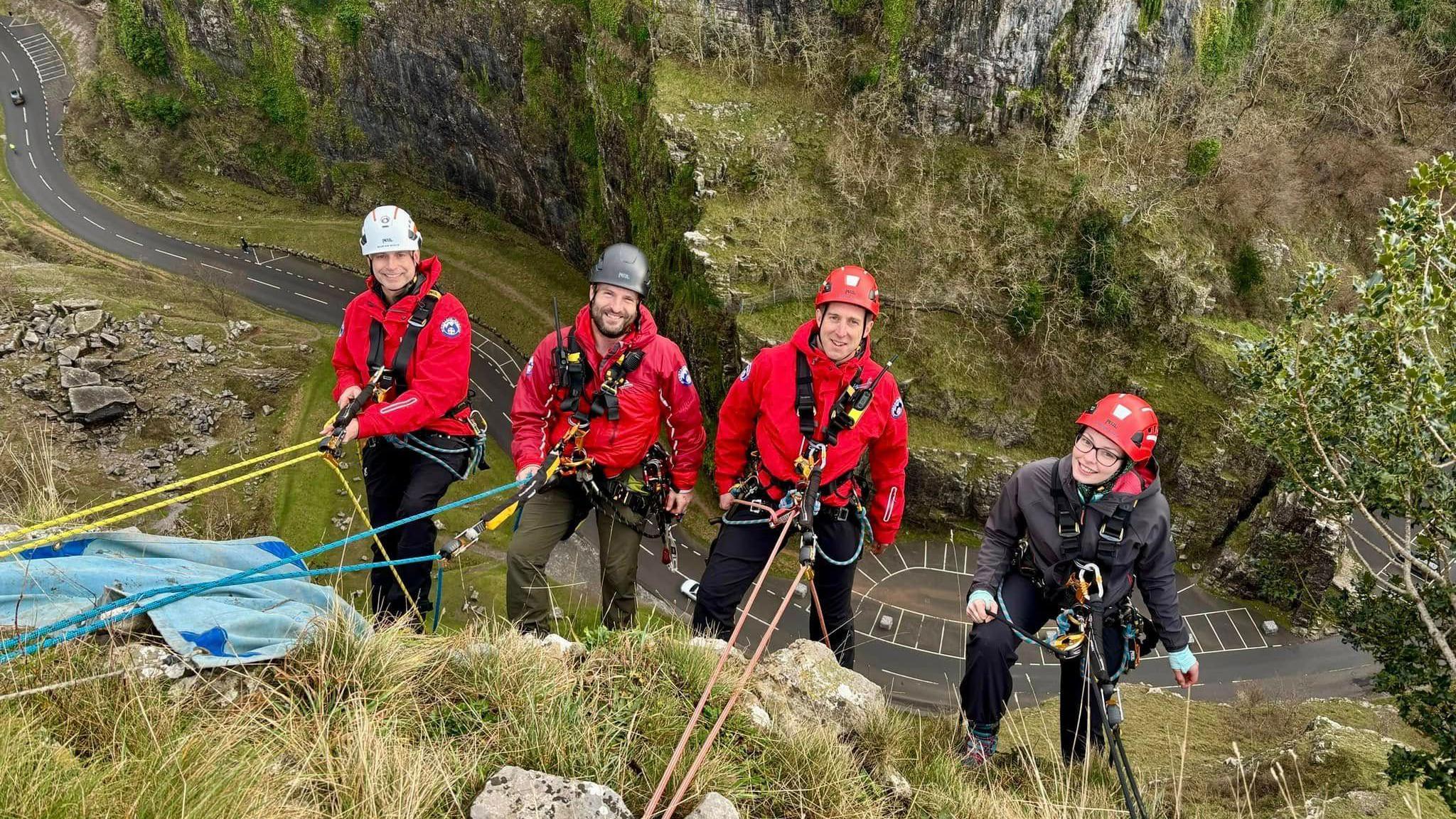 Four people are on the edge of Cheddar Gorge, fastened to ropes. They are wearing red jackets and red helmets.