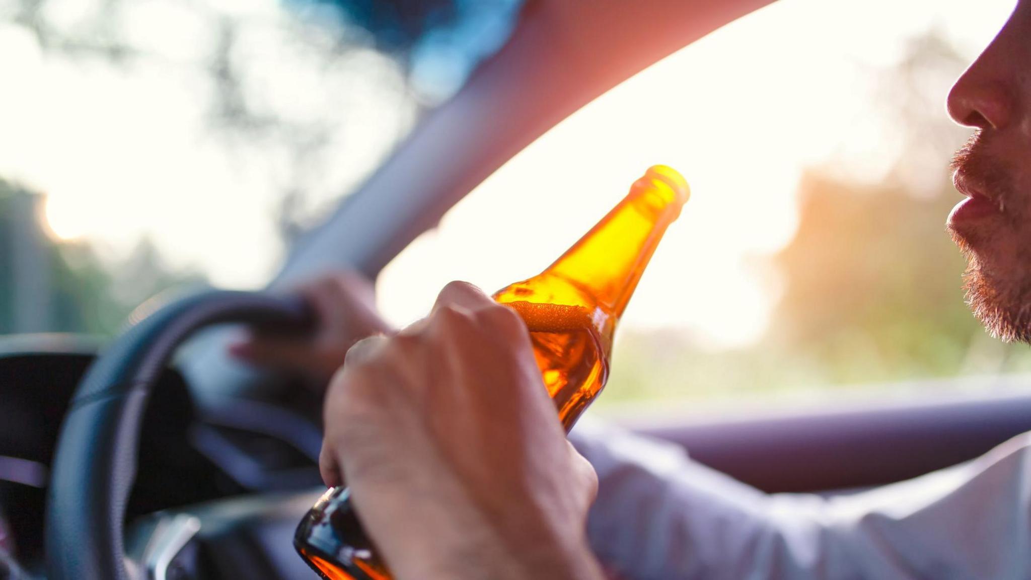 A stock image of an anonymous man sat in the driver's seat of a car with one hand on the wheel and the other holding a brown beer bottle