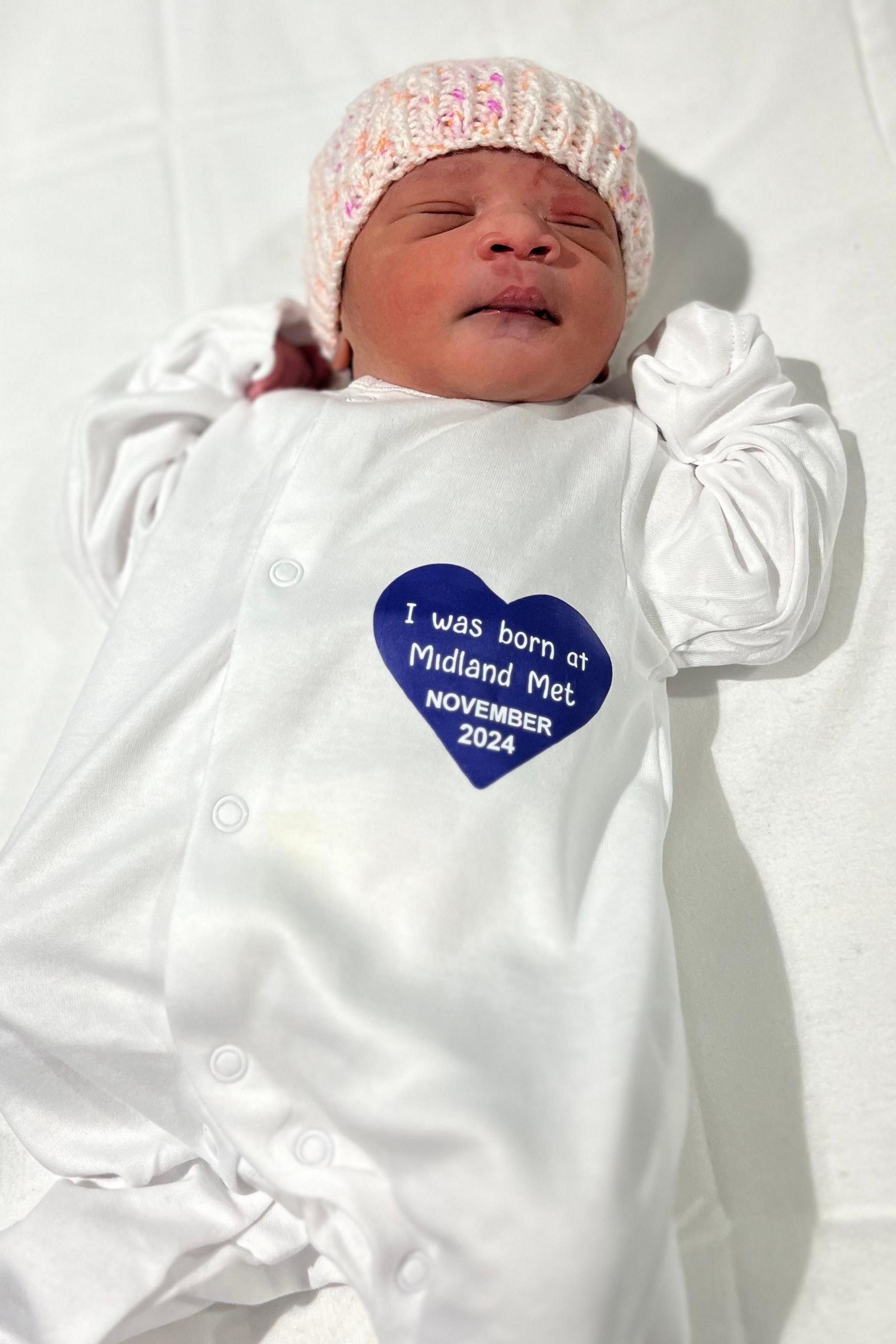 A baby in a white babygrow and a knitted hat, lies on a white bedsheet. The babygrow has a blue heart with the words "I was born at Midland Met November 2024" written inside it.