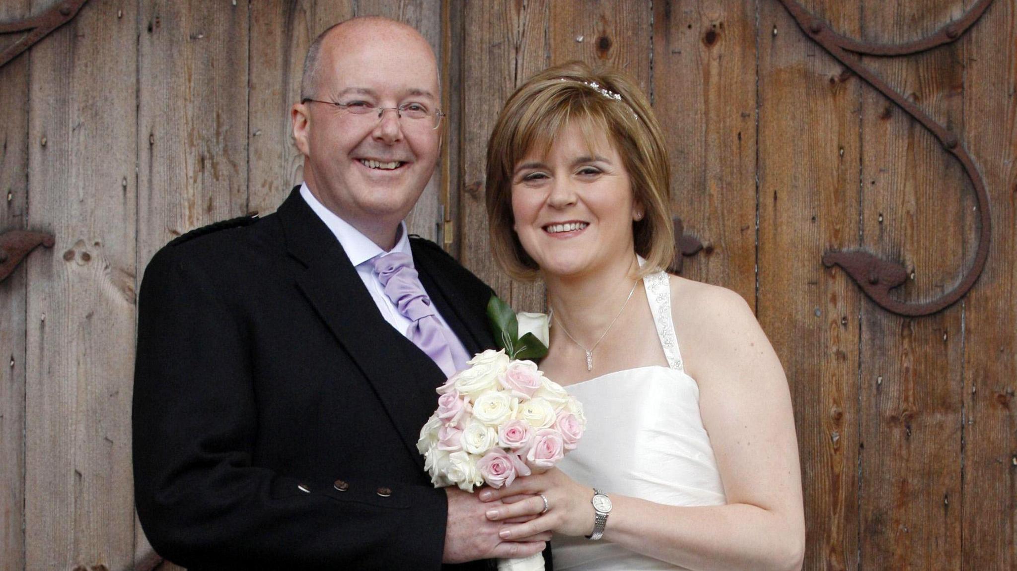 Peter Murrell on the left wearing a black jacket over a white shirt and a lilac tie. He is smiling at the camera. He is bald on the top of his head with grey hair around the side. He has glasses on his face and is holding a bouquet of white and pink flowers. Nicola Sturgeon is on the left. She has shoulder length dark orange hair and is smiling at the camera. She is also holding the bouquet. She is wearing a wedding dress. Both are standing in front of a large wooden door.