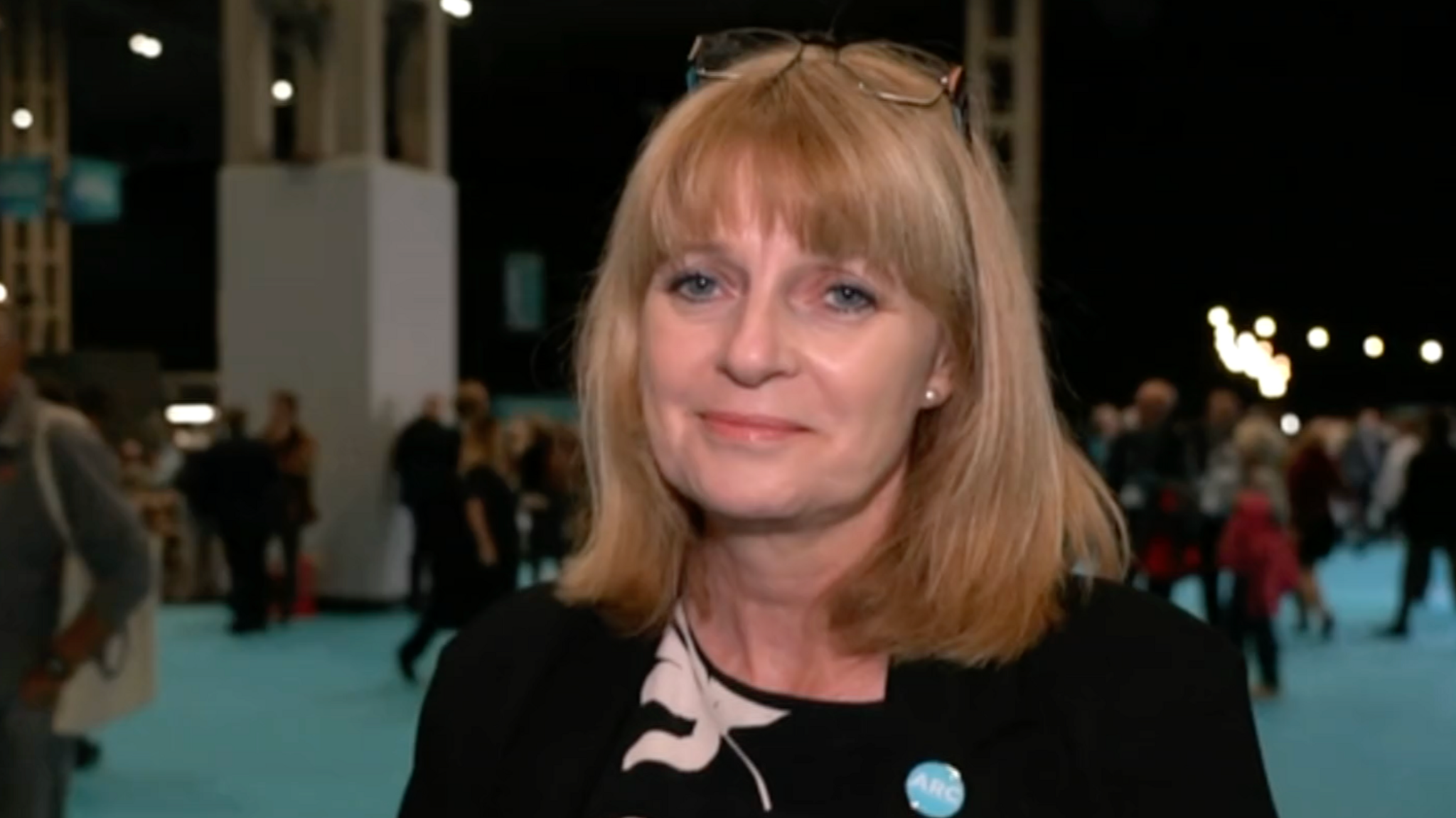Jackie Gardiner has blonde shoulder length hair and is wearing a black top. She is standing in a large room with people walking on a green carpet behind her.