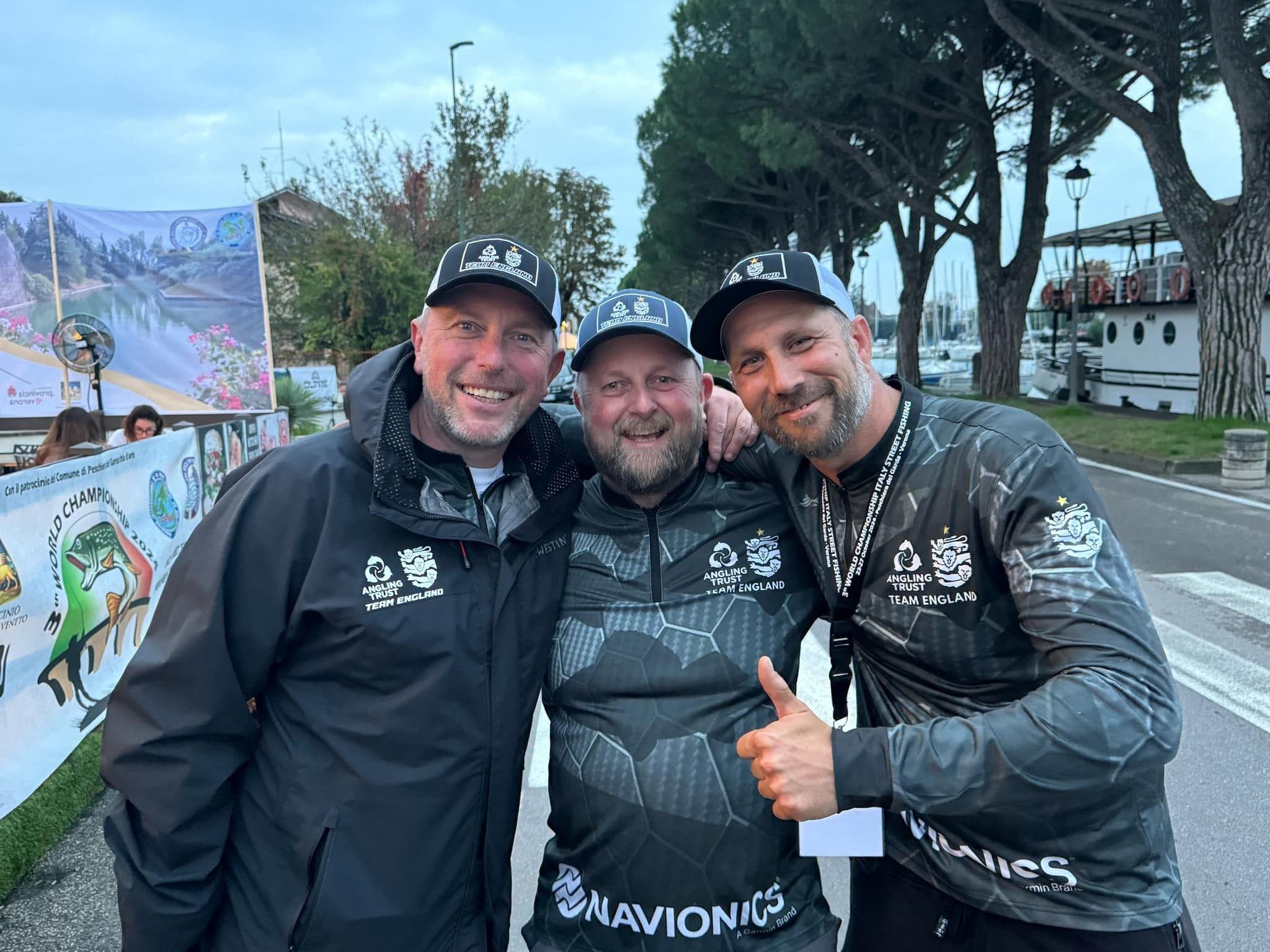 Three main hold each other and smile at the camera. They are wearing baseball caps. The man on the right is holding his thumb up. They are on an urban street.