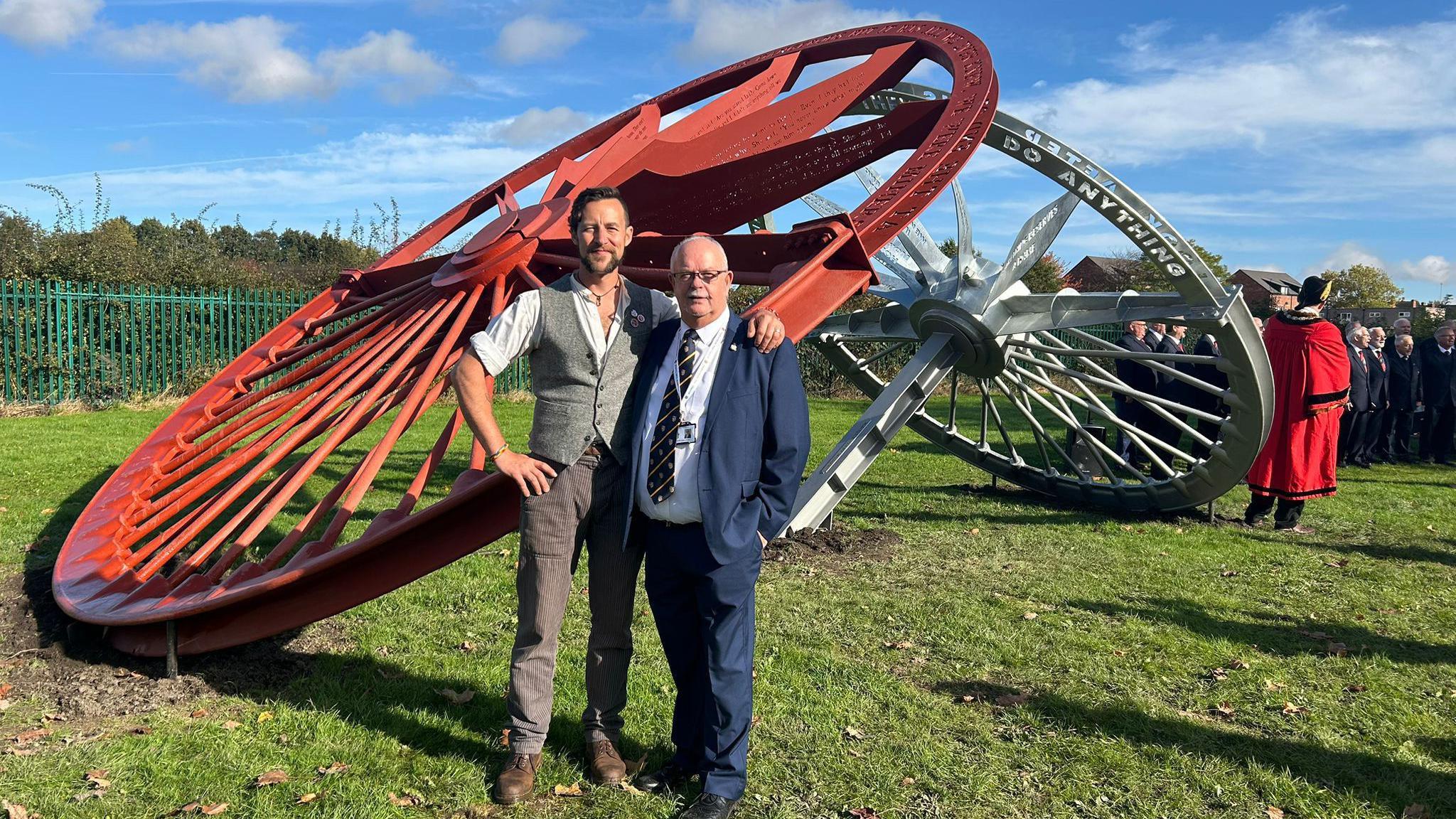 Artist Luke Perry with Councillor Stephen Vickers next to the pit wheel sculpture. The sculpture, with one red pit wheel and one in silver, towers over them. In the background there's a man in mayoral dress and a male voice choir.  