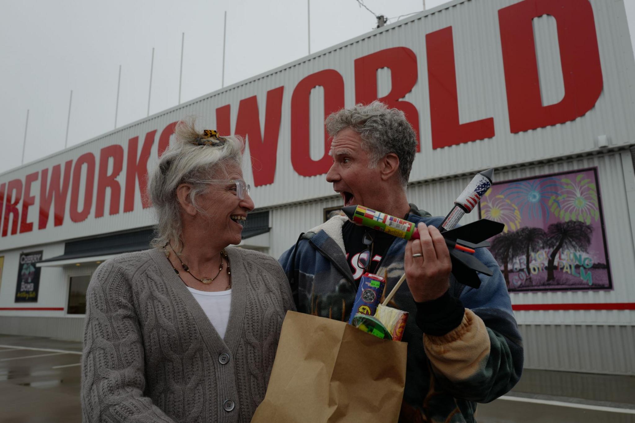 Harper Steele,  wearing a white top and taupe cardigan, holding a bag filled with fireworks alongside Will Ferrell, who is holding some firework rockets, wearing a blue jacket.