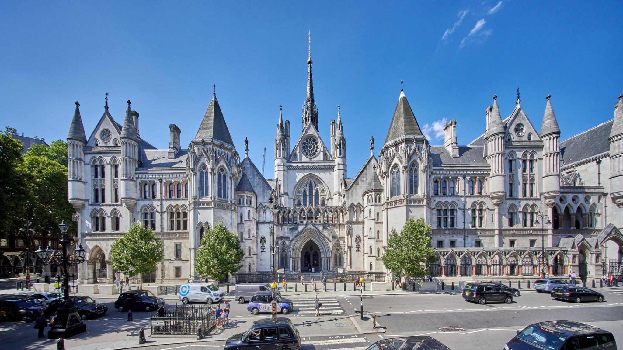 An exterior view of the Royal Courts of Justice, showing its Gothic Revival spires and turrets against a bright blue sky, with cars passing on the road outside