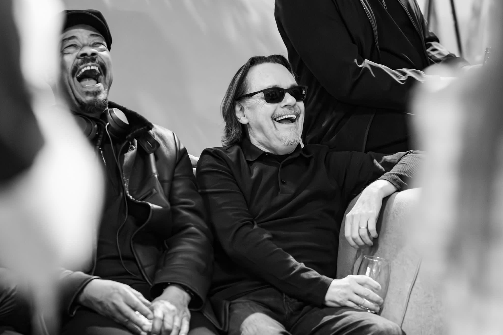 Earl Falconer sits to the left of his band-mates in a black and white shot where his head is tilted back as he enjoys a big laugh