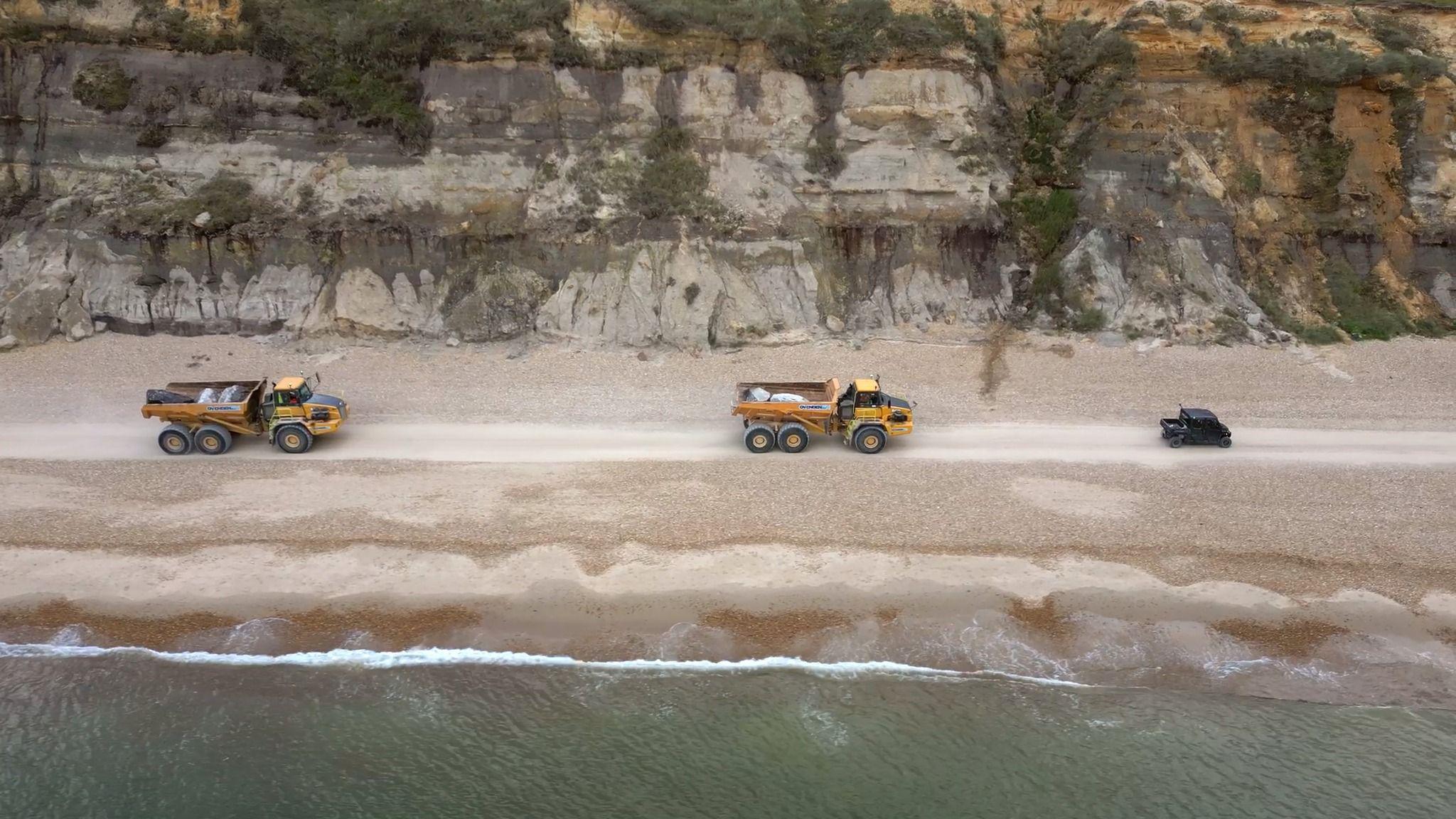 Two yellow lorries on a beach and a smaller black vehicle. The picture was taken from a bird eye's view. It is an overcast day.