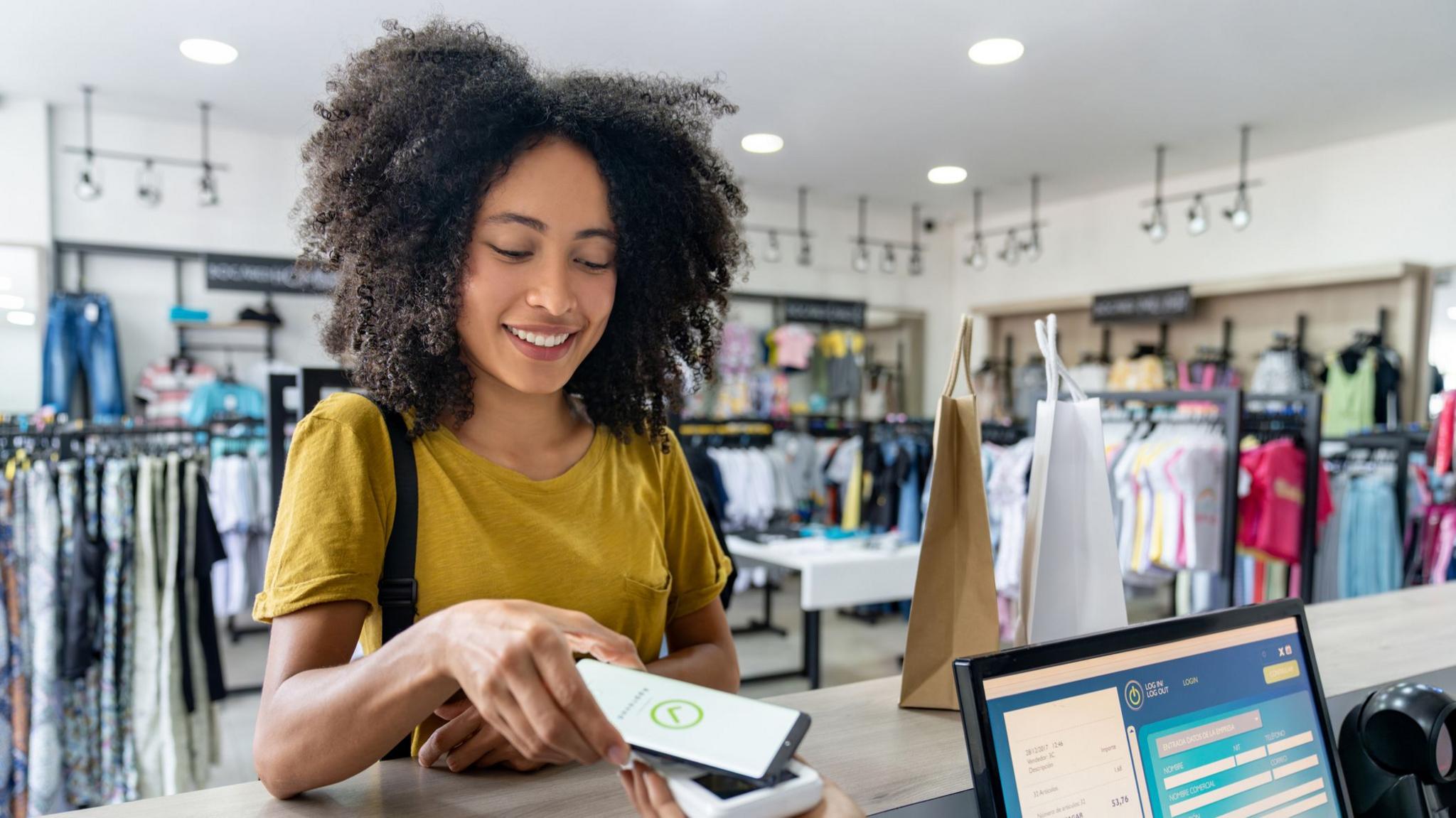 Young woman with dark, curly hair wearing a mustard yellow t-shirt pays at a counter in a clothes shop using her smartphone  