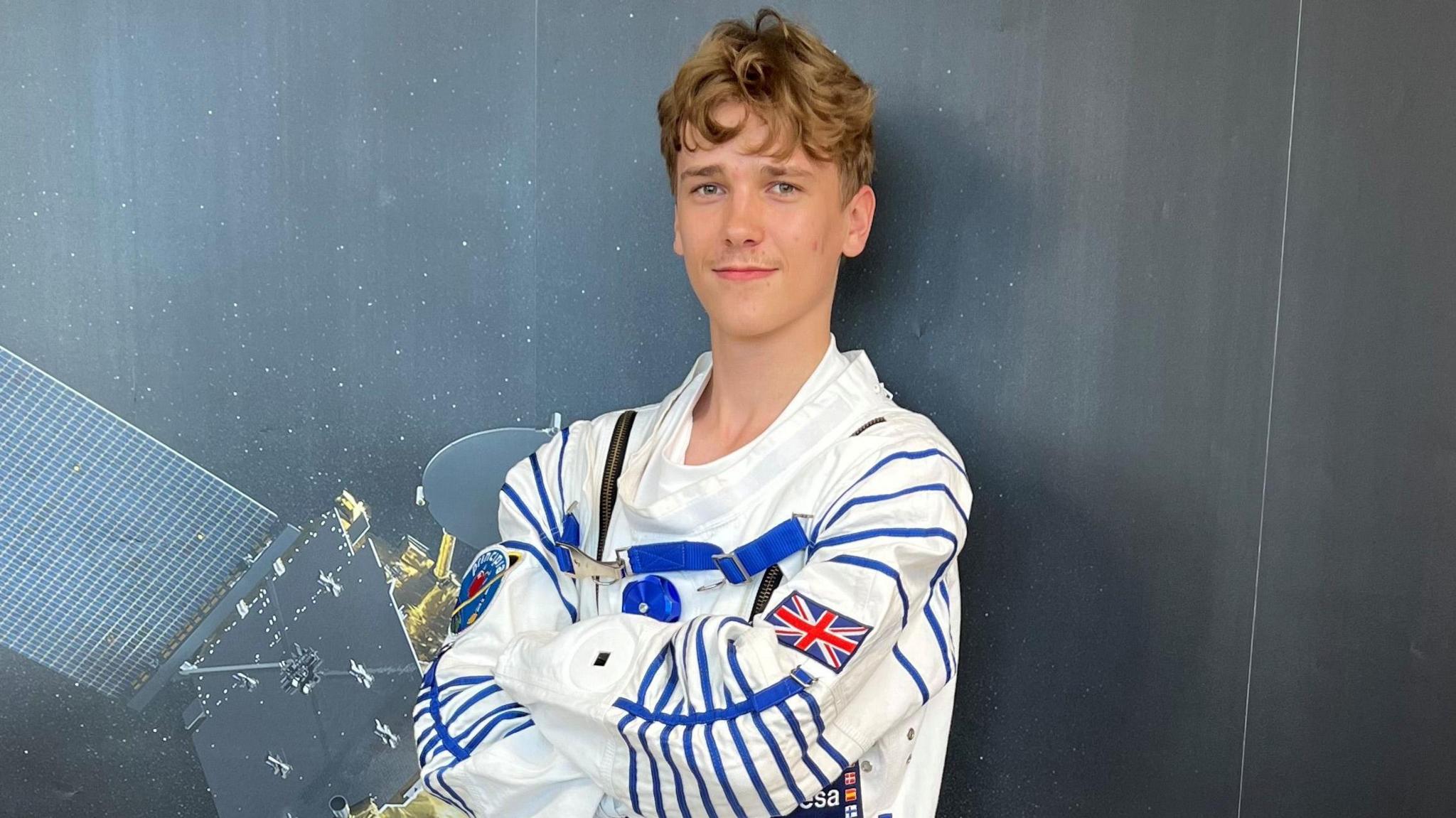 Maxim is wearing a space suit and posing behind a satellite print. 