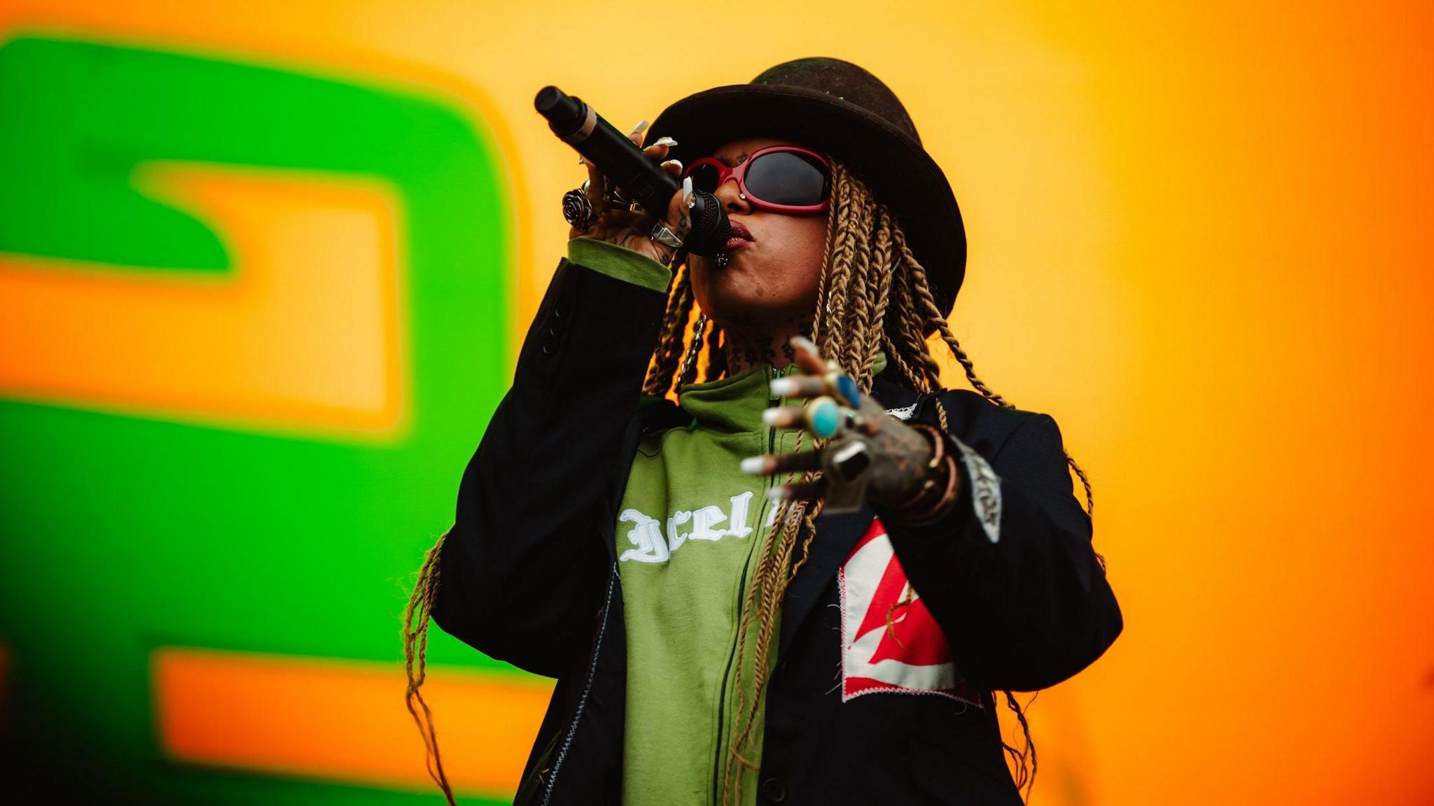 A woman performs on stage at Forwards Festival in Bristol. She is wearing a dark jacket and a hat and is singing into a microphone