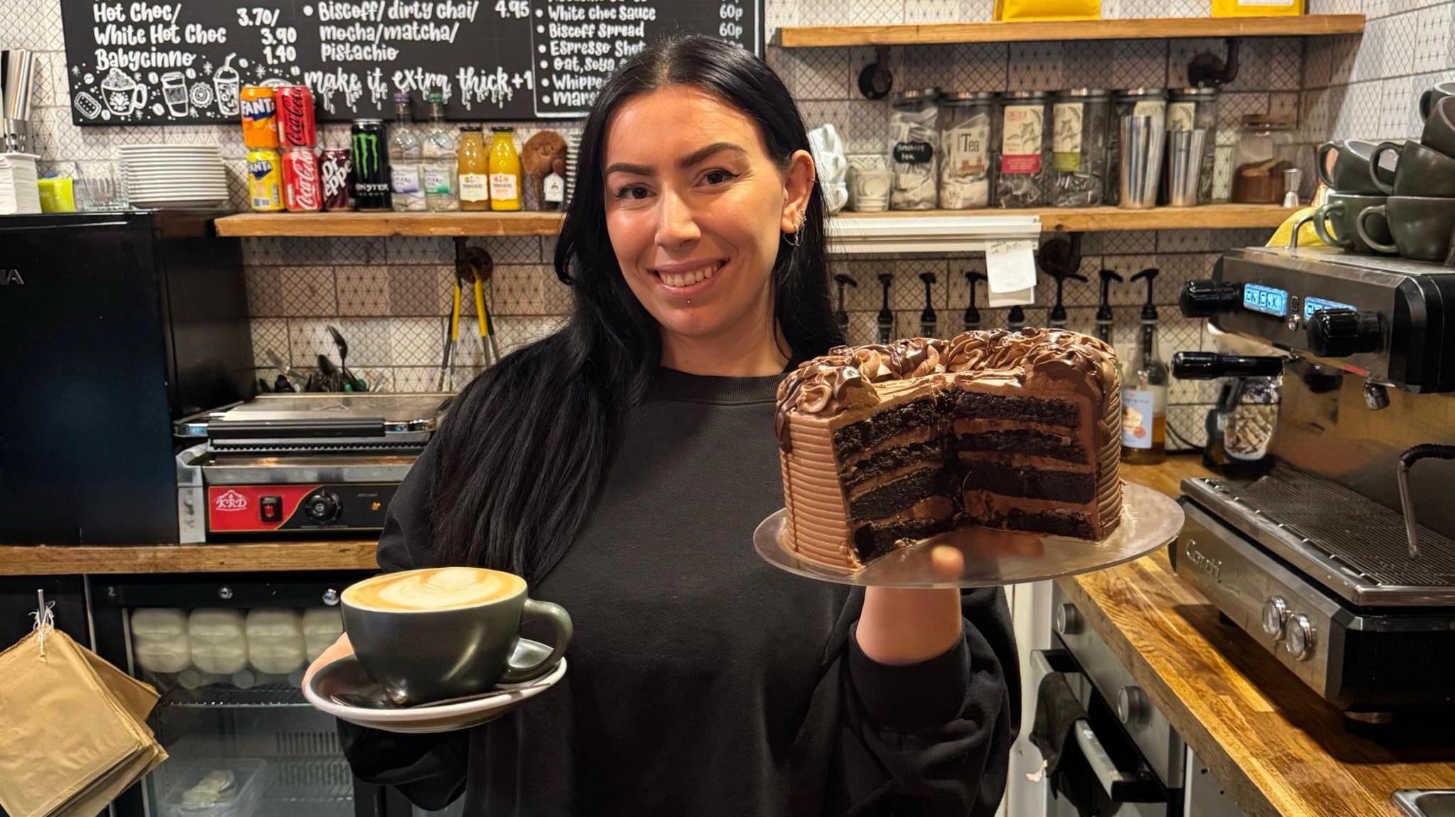 A woman with long black hair and an oversized black jumper is smiling into the camera whilst holding up a big chocolate cake which has been cut in half in one hand and in the other she is holding a black cup of latte in a saucer. Behind her is a coffee machine, a fridge, various drinks and teabags and a board with a menu on it written in white.