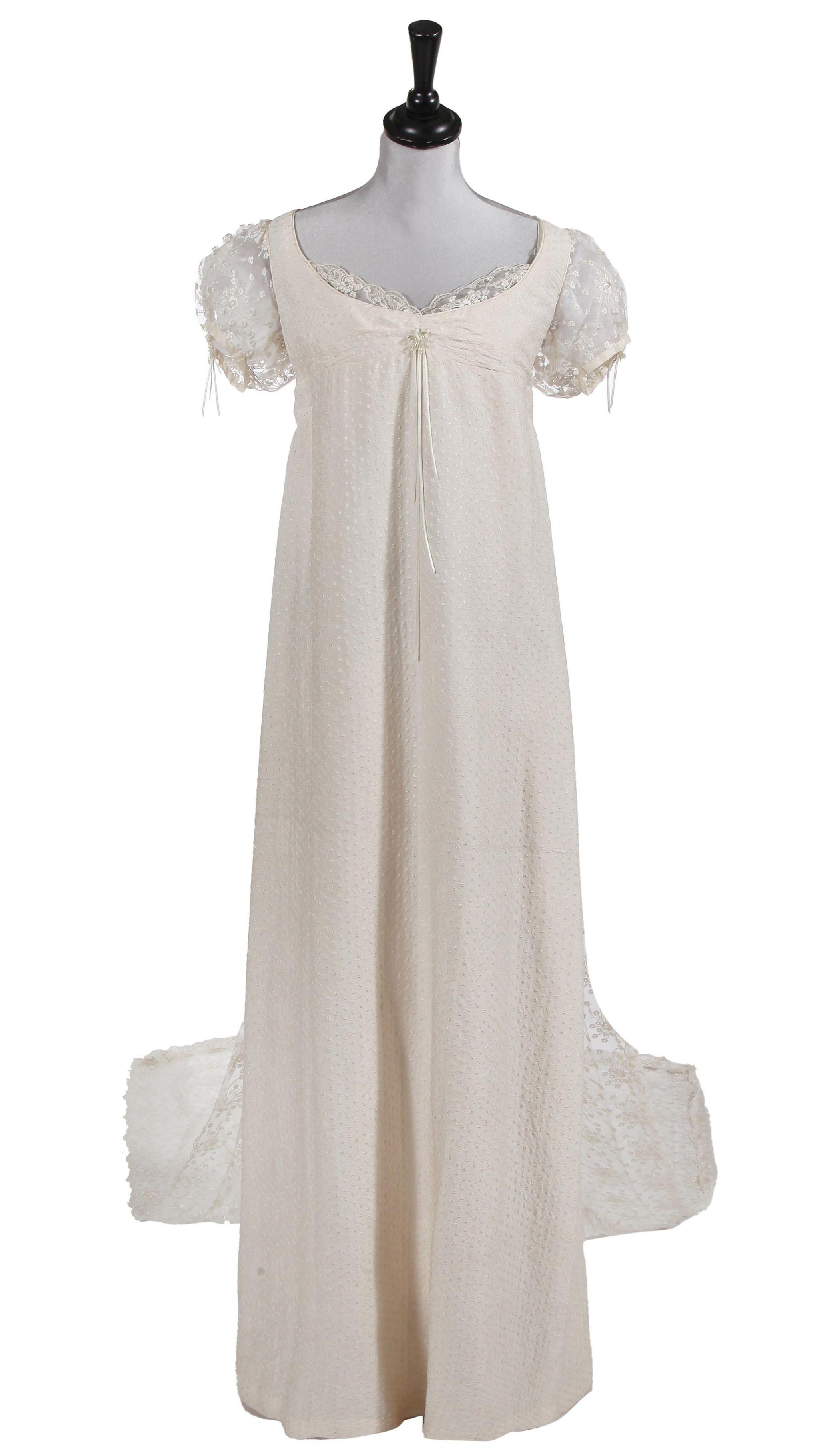 A circa 1810 style wedding gown of textured white cotton with overall broderie anglaise embroidery, a bodice with a scoop neck trimmed with lace, high empire waist, and faux pearl and ribbon detail to centre front, with short puff lace sleeves, also featuring a white underslip and a white lace train.