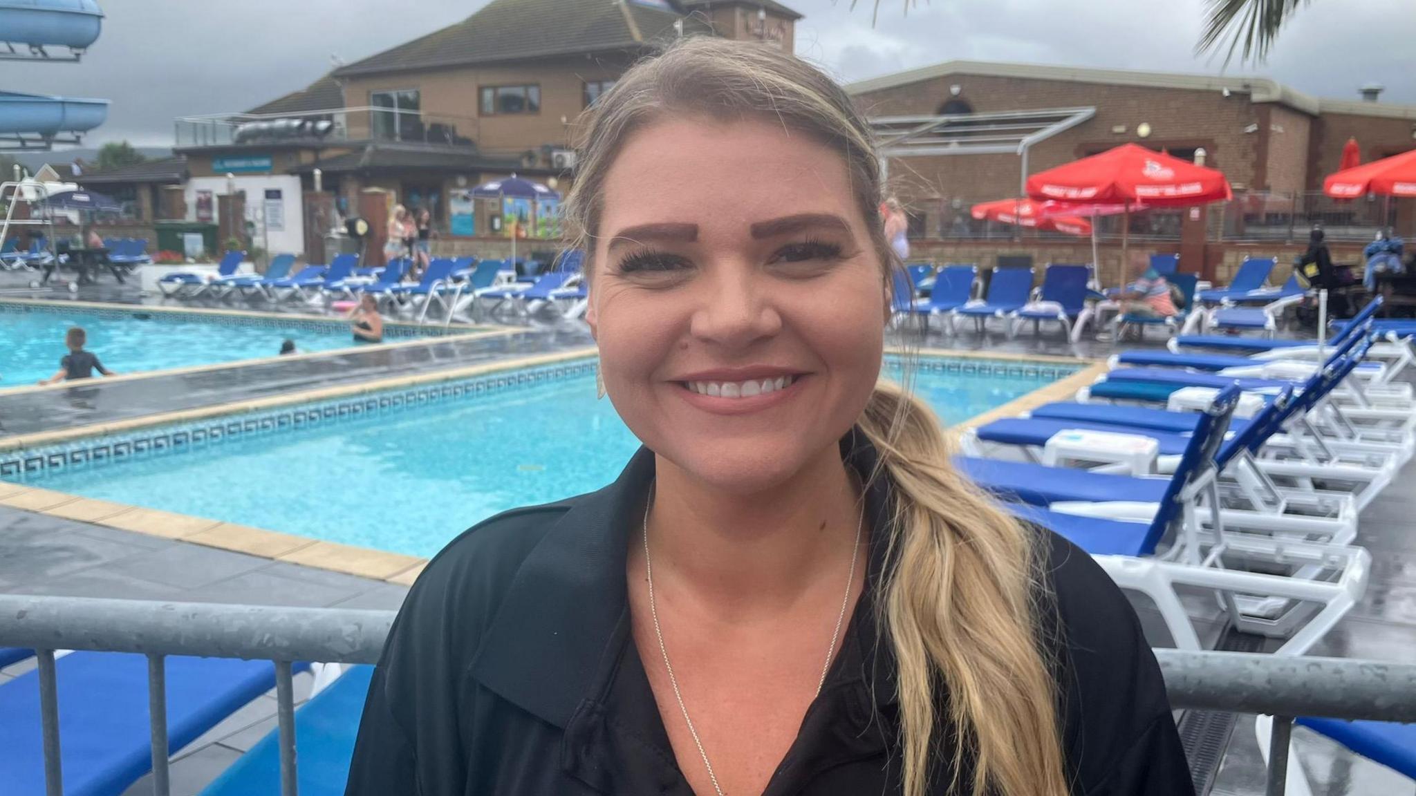 Rhian Hughes stood in front of an outdoor swimming pool and smiling at the camera. The swimming pool is surrounded by blue and white sun loungers and part of a large water slide can be seen to the left. There are also a number of bright red sun umbrellas.