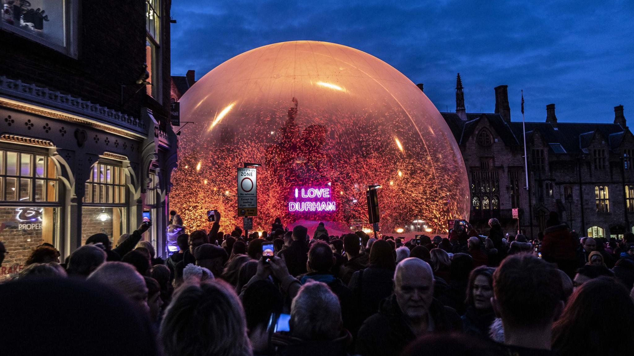 A monument in Durham Market Square is covered in a snowball like structure with snowflakes flying in it. A neon pink sign in front of the monument reads I LOVE DURHAM. A big crowd of people has surrounded the structure.