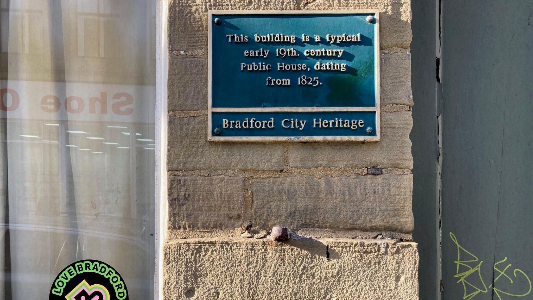 A close-up of a blue plaque with white lettering fixed to the outside wall of the pub which reads: "This building is a typical example of an early nineteenth century public house, dating from eighteen twenty five." Underneath that also in white letter are the words Bradford City Heritage. 