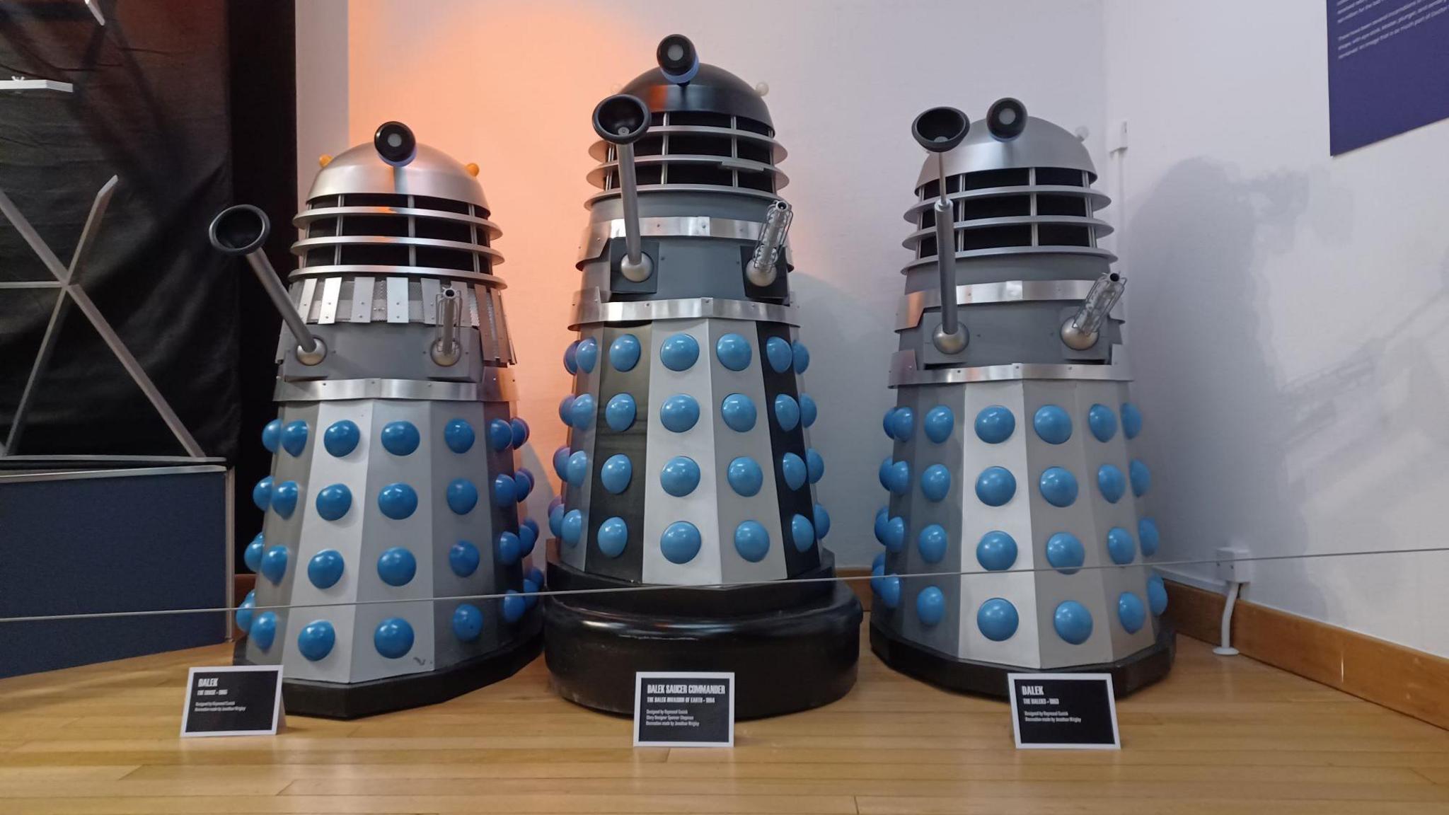 A group of three prop Daleks, conical robotic alien creatures, from the BBC science-fiction series Doctor Who, on display as museum exhibits against a white background. The two on either side are silver with blue trim, while the one in the centre stands slightly taller on a large black fender, with black stripes and a black dome section at the top in addition to the silver and blue of the others.