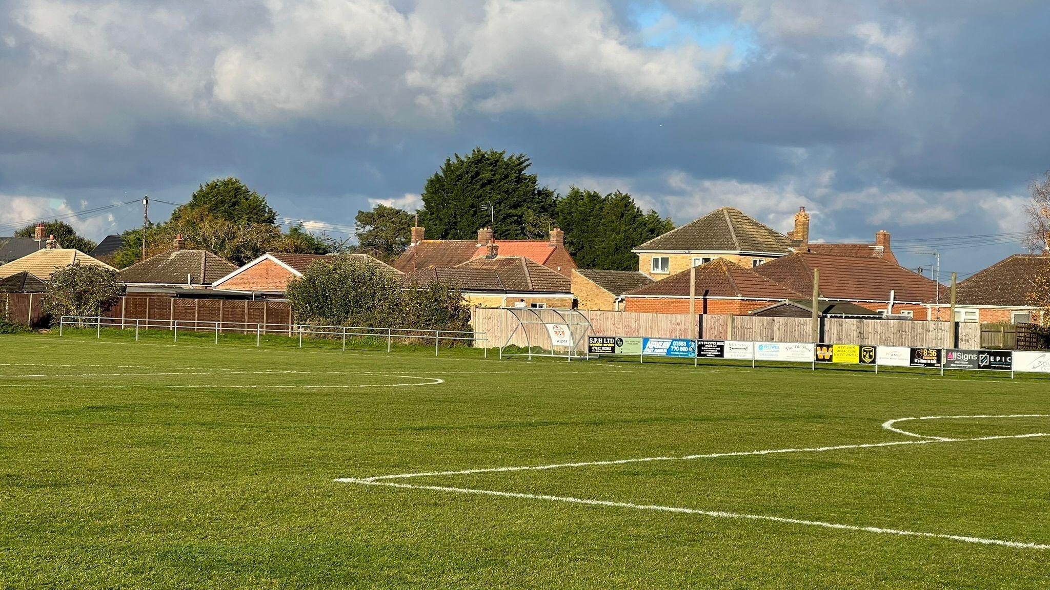A grass football pitch is shown with houses in the background beyond a fence.