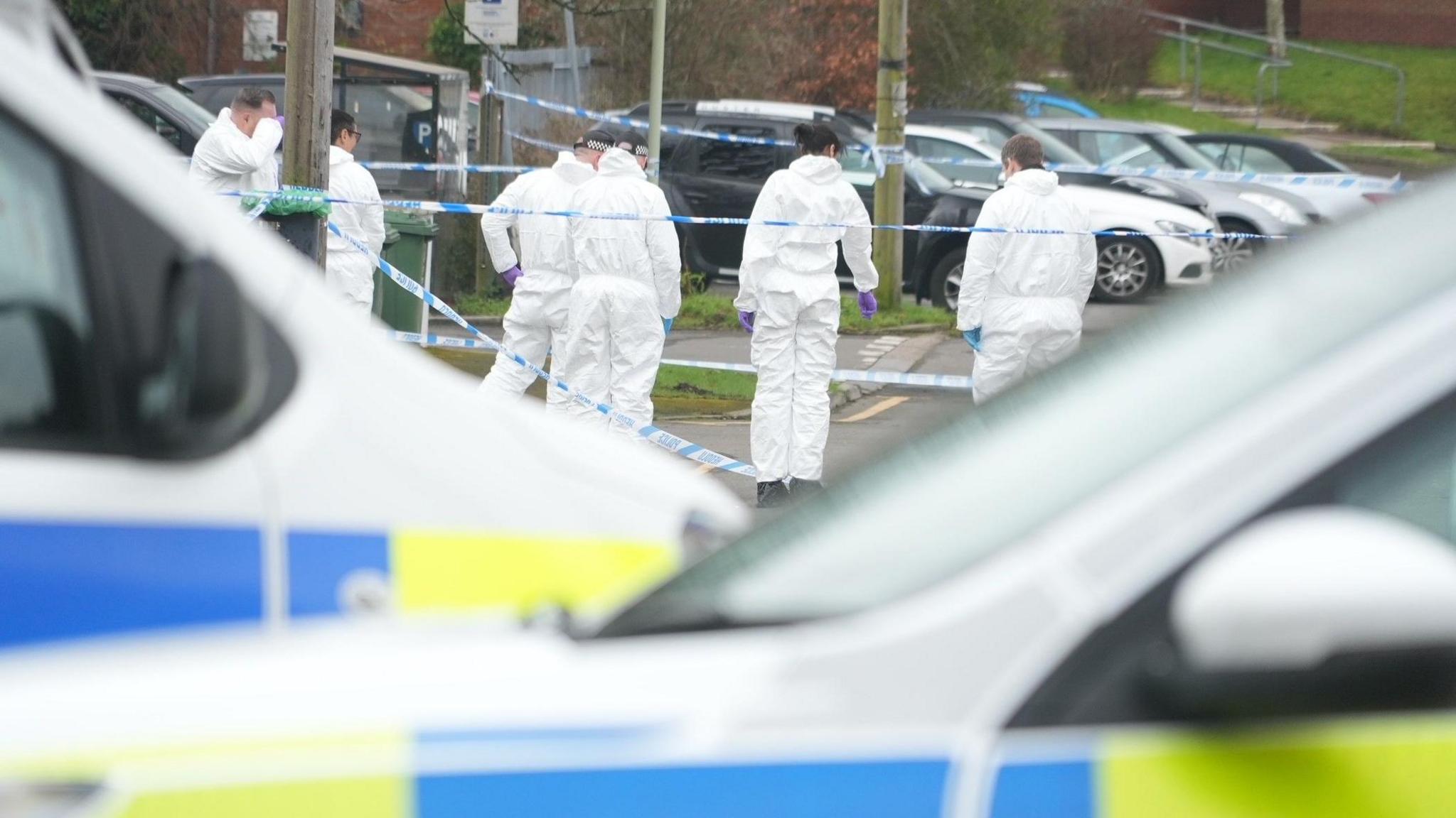 Several investigators in white overalls survey the scene behind a police cordon and police vehicles in the foreground