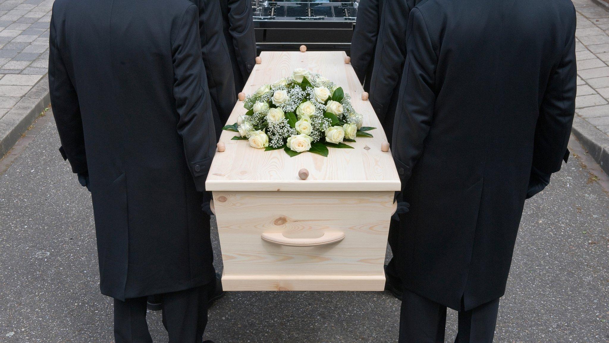 Pallbearers carry an MDF casket adorned with flowers at a funeral ceremony