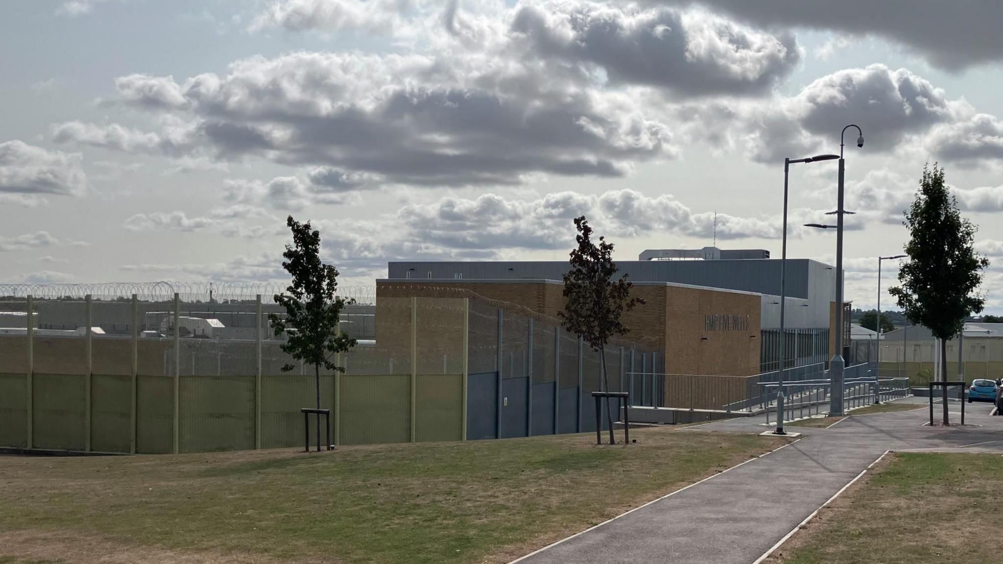 An external view of HMP Five Wells, with perimeter fencing, trees and grassy areas outside, and the modern prison buildings visible