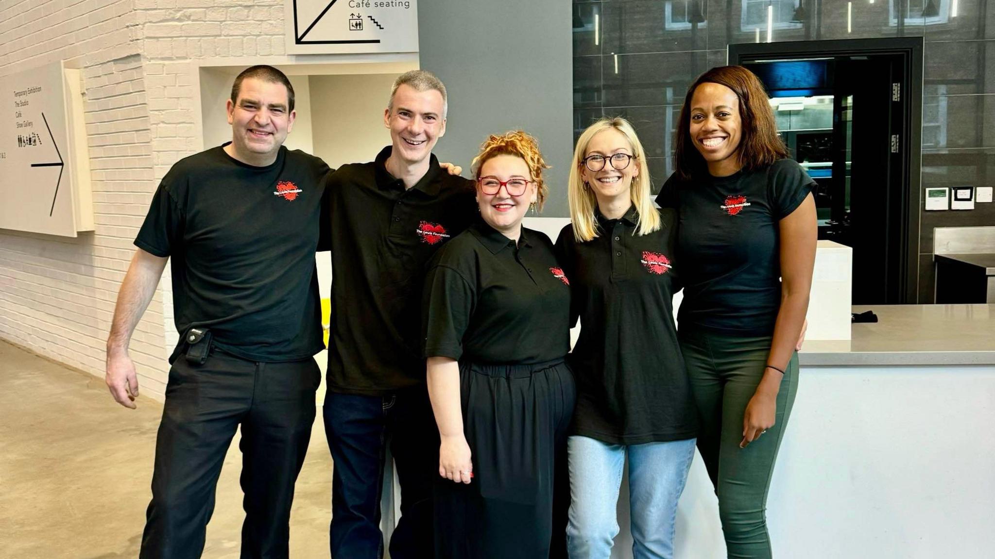 The Lewis Foundation team -  three women and two men stand smiling in the empty cafe, looking at the camera. They are all wearing the black,  branded "Lewis Foundation" T-shirts.