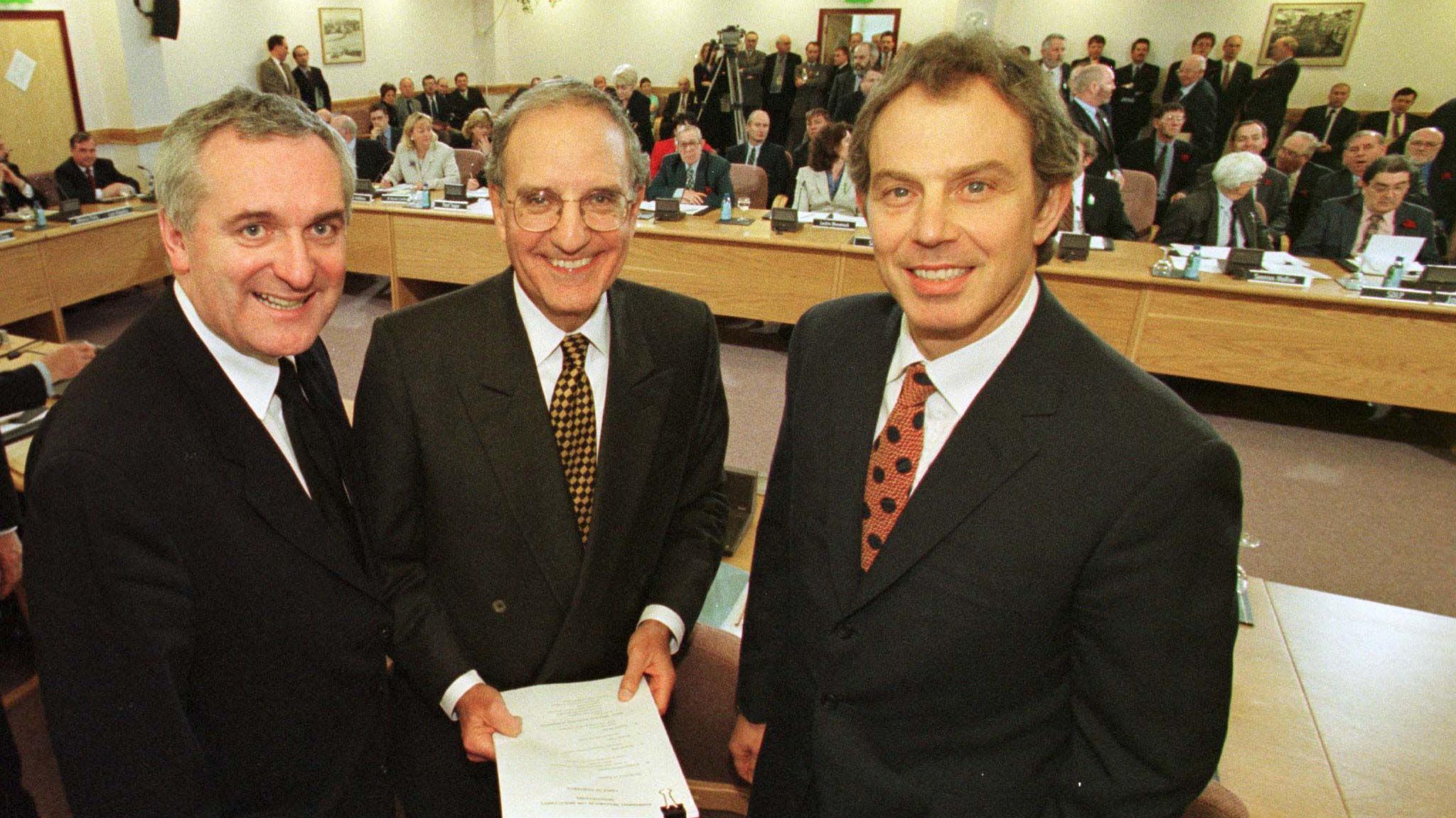 Three men stand in front of a table in a room full of other politicians. They are all smiling, Bertie Ahern is on the far left. He has short, grey, receding hair. In the middle is George Mitchell. He also has short grey hair and is balding. He is wearing glasses and is holding a big sheaf of papers. Tony Blair is on the right of the picture. He has short brown hair. All three are wearing dark coloured suits with white shirts and multi-coloured ties.